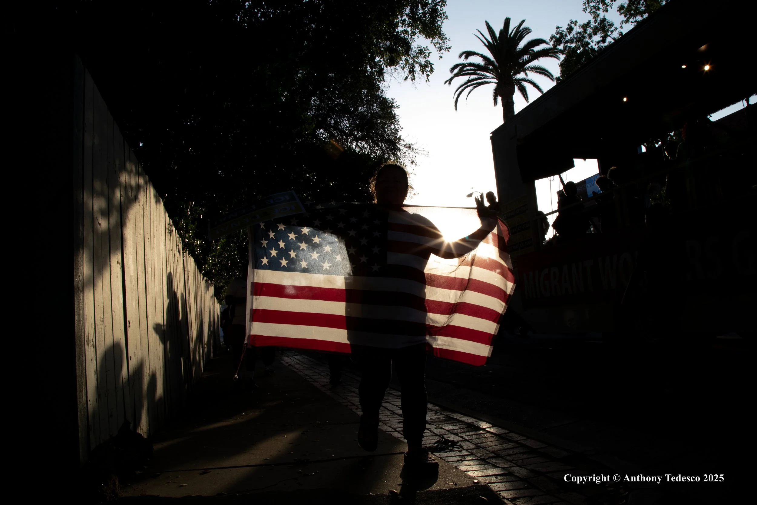 Person holding an American flag in front of the sun, with shadows cast on a wooden fence and a street scene in the background, including palm trees and people on a boat or float.