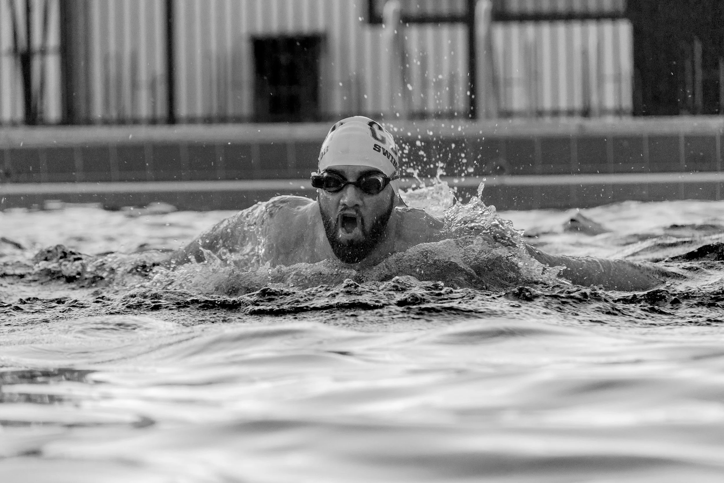 A swimmer in a pool wearing a swimming cap and goggles, actively swimming front crawl with water splashing around.