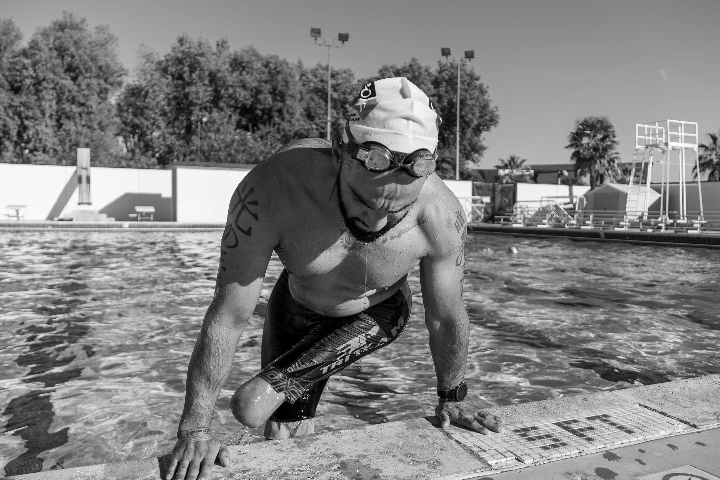 A male swimmer emerging from a swimming pool, wearing a swim cap and goggles, with tattoos on his arm and sitting on the pool edge.