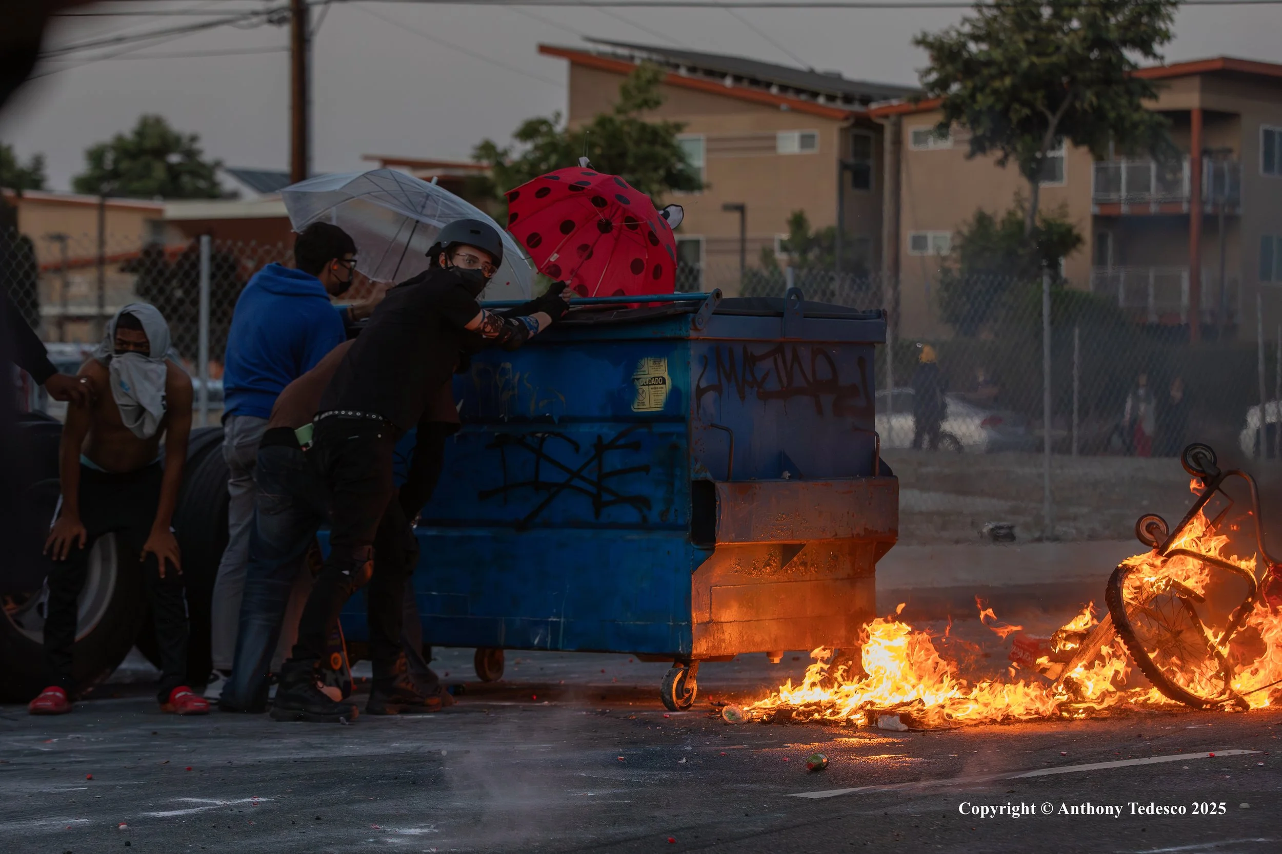 People wearing masks and helmets pushing a blue dumpster as a bicycle behind it catches fire on the ground during a protest or riot, with infrastructure and buildings in the background.