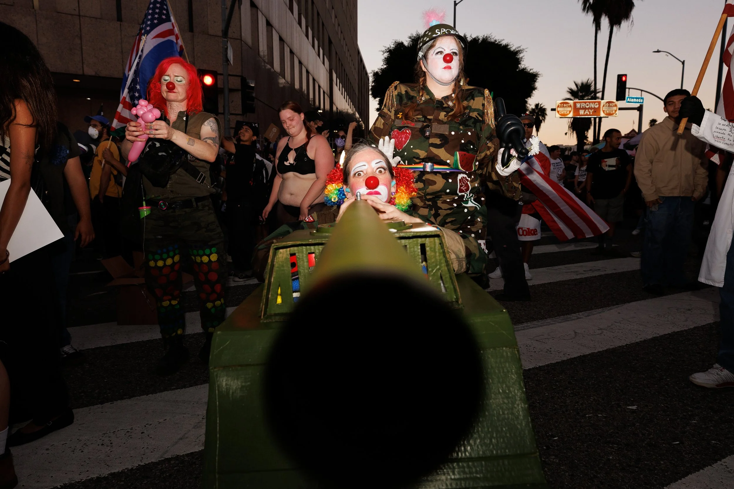 Clowns dressed in military camouflage costumes with red noses at a street protest, one is operating a cannon, others holding flags and balloons, with a crowd and city street in the background during dusk.