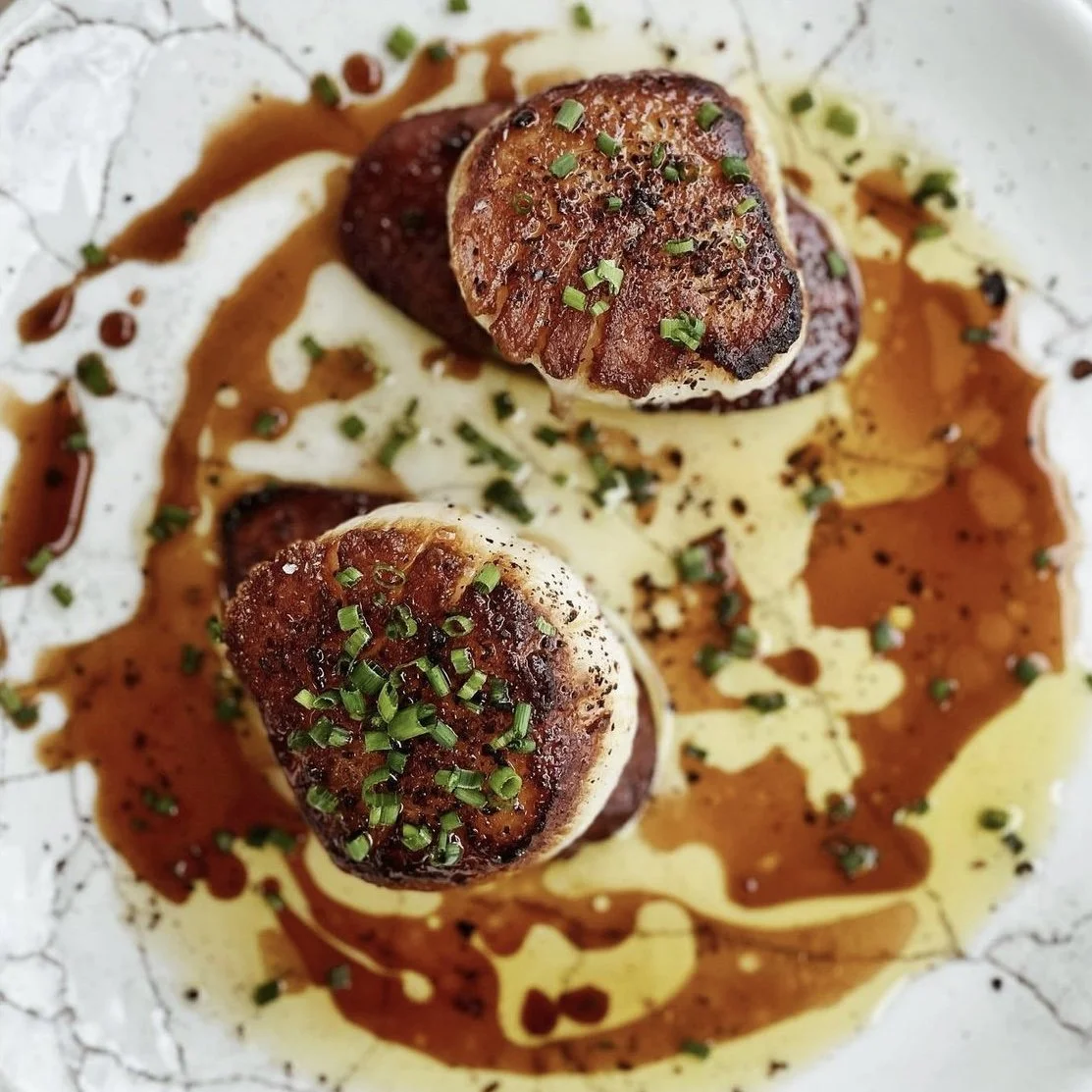 Close-up of seared scallops garnished with chopped chives, served on a white plate with sauce drizzled around.