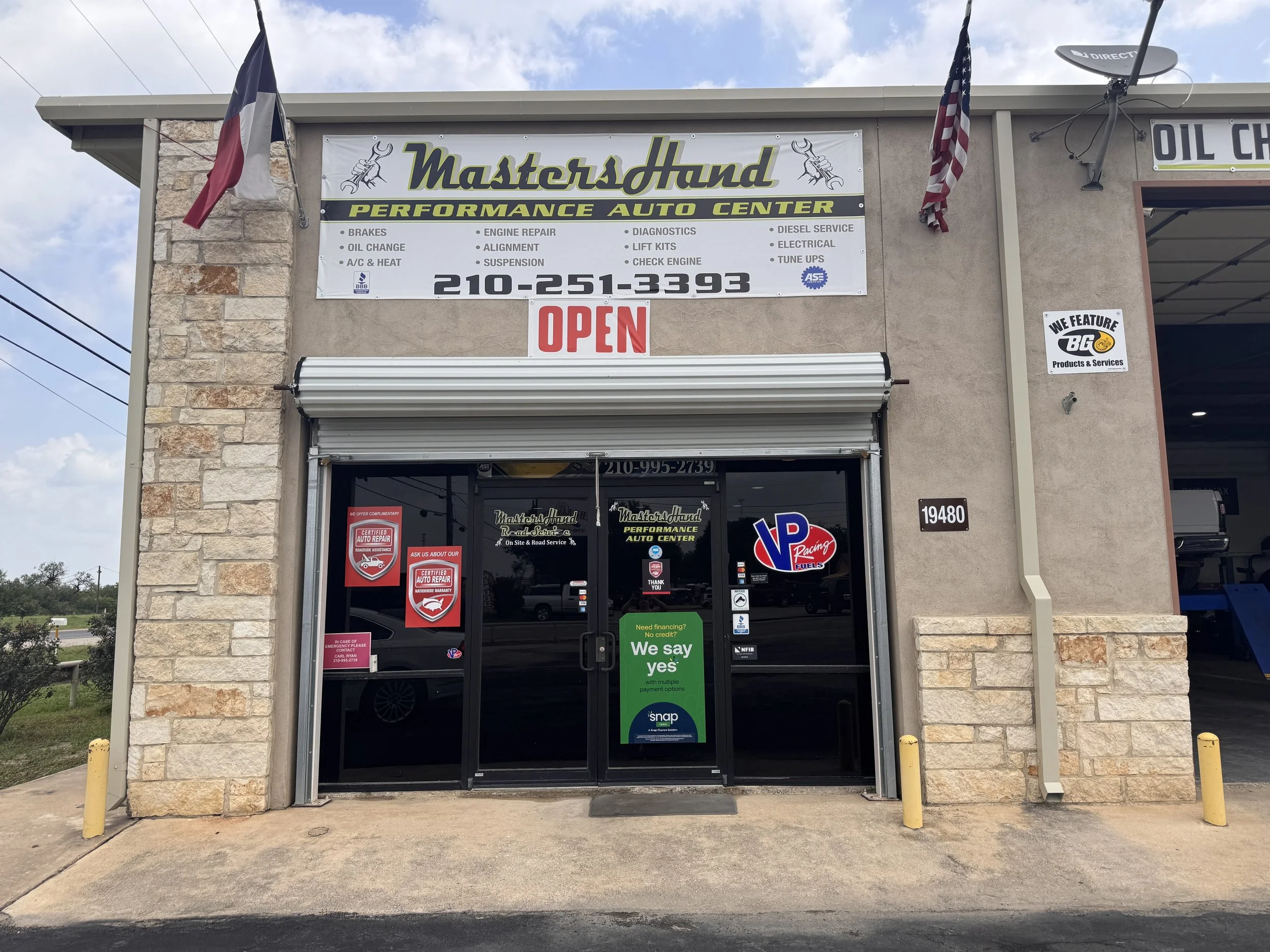 The storefront of Masters Hand Performance Auto Center with a sign displaying services, the word 'OPEN' in red, and various stickers on the door.