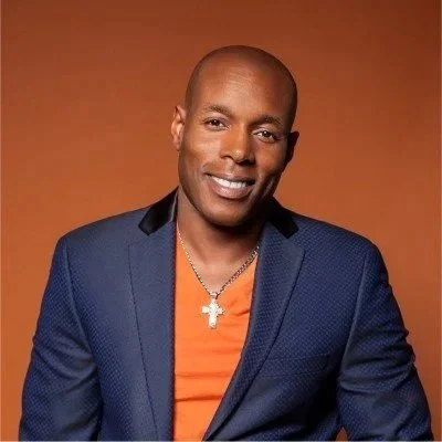 Portrait of a man wearing a blue blazer, orange shirt, and a silver cross necklace, smiling against a brown background.