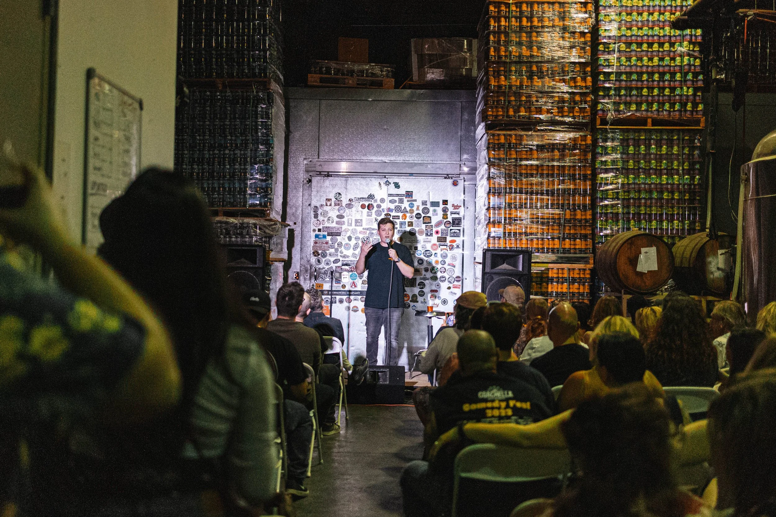 A comedian performs on stage during a stand-up comedy show at a bar or brewery, with audience members seated and watching. The backdrop features stacked beer cans and large barrels.