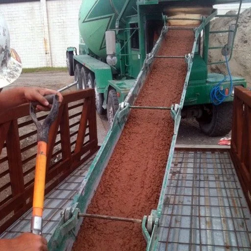 A concrete mixer truck pouring wet concrete onto a metal ramp, with a person holding a rake on the left side.