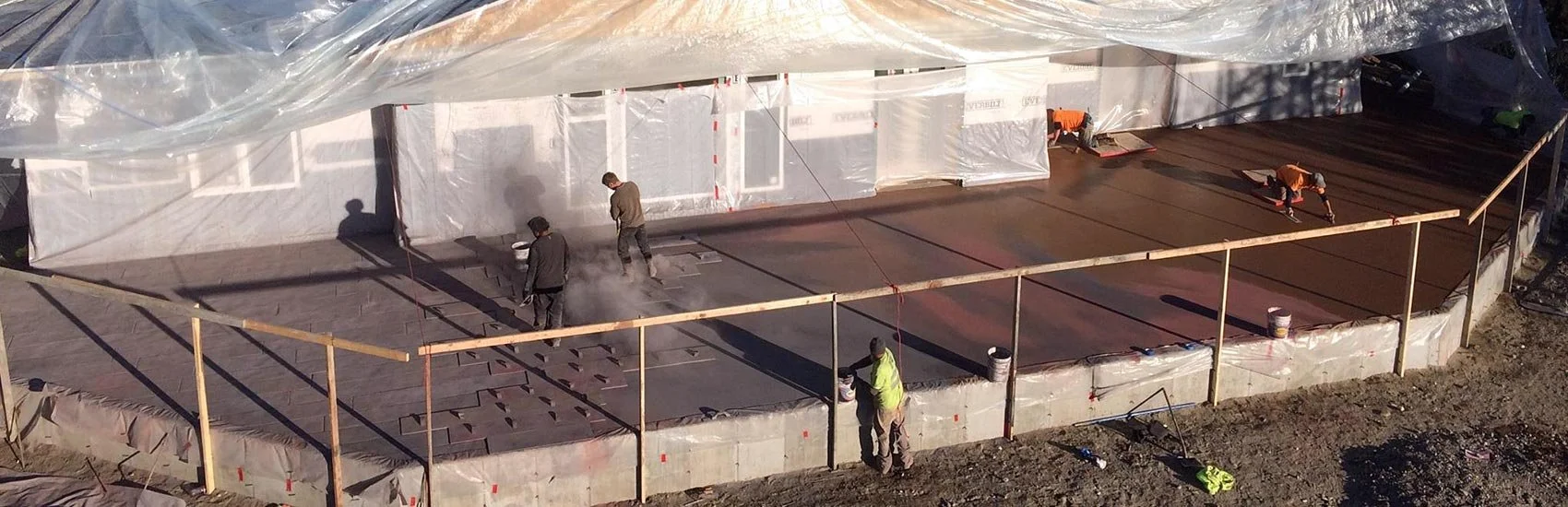 Construction workers are pouring and leveling concrete on a building's foundation, surrounded by scaffolding and plastic sheeting.