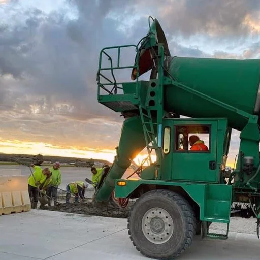 Construction workers pouring concrete at a job site during sunset, with a large green cement mixer truck in the foreground.