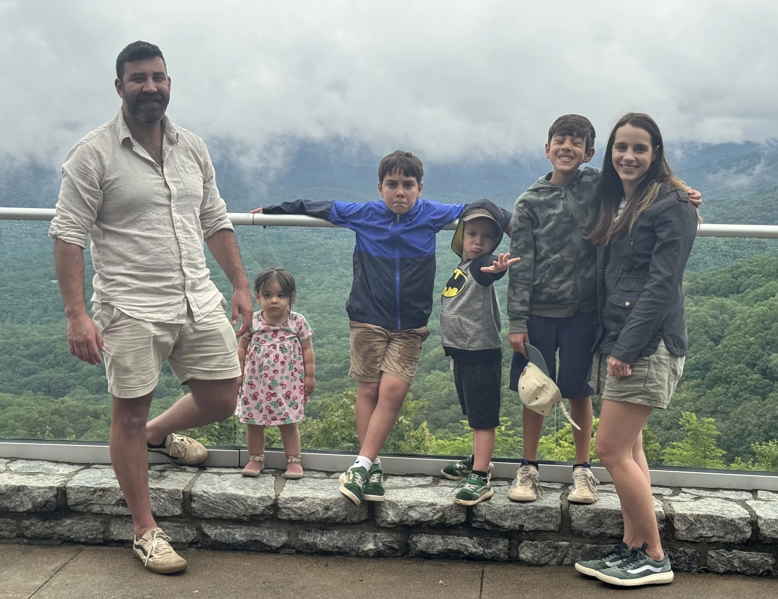 Family of six posing on a balcony with a mountain landscape in the background, under cloudy weather.