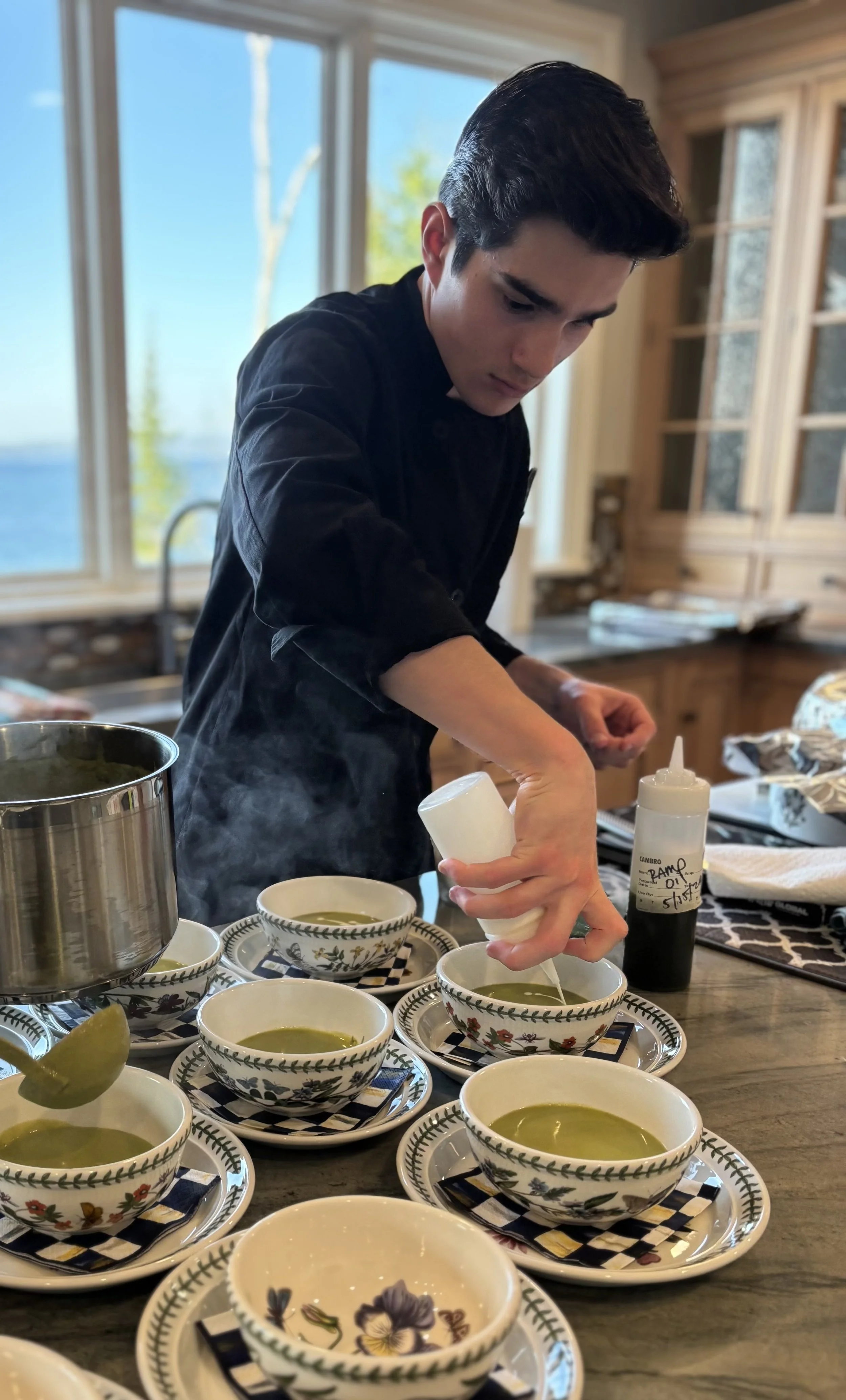 A young man in a black shirt preparing tea in a kitchen with large windows showing trees outside. Several bowls of green tea and matching saucers with floral patterns are on the table.