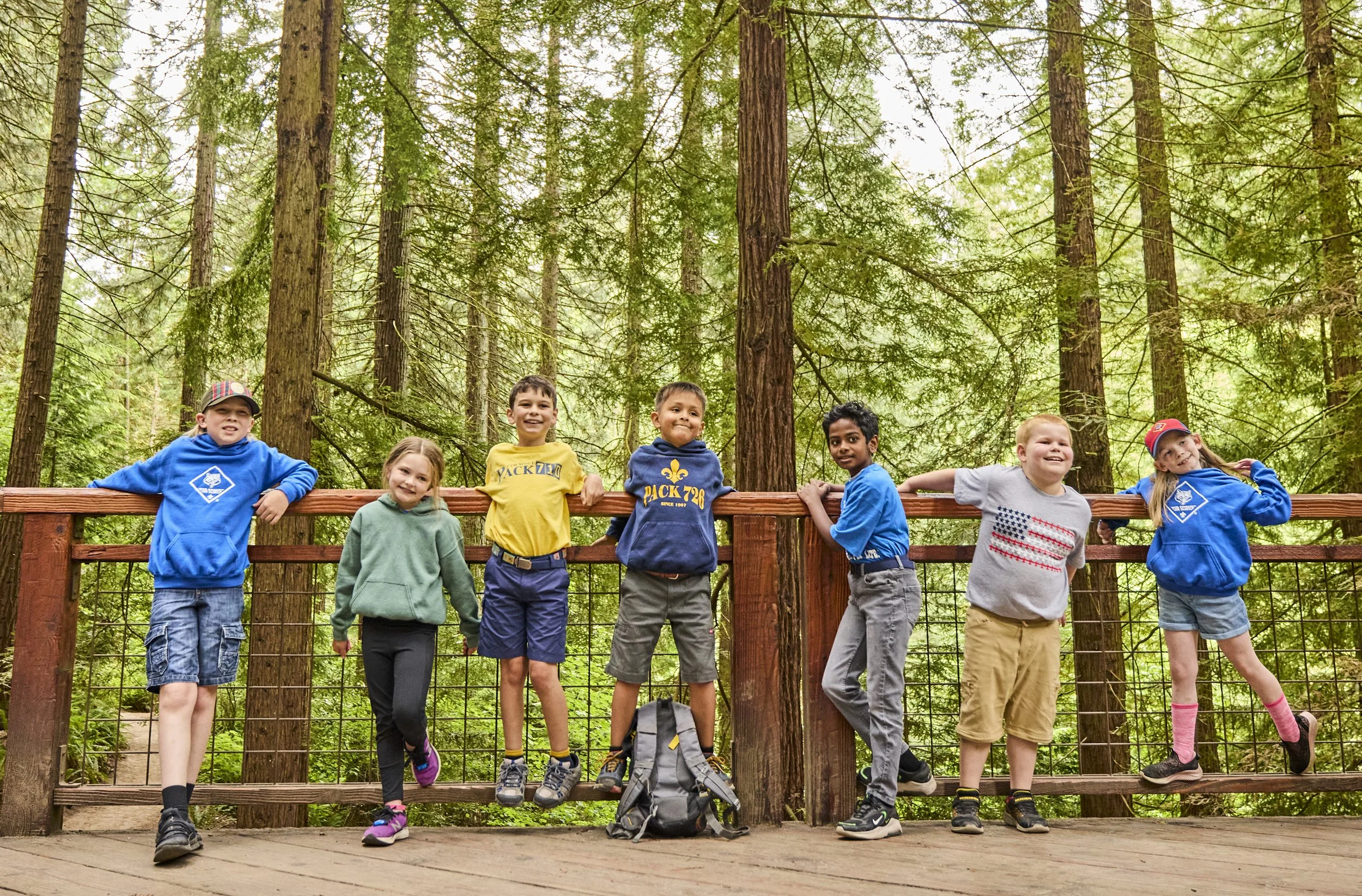 Group of seven Cub Scout children standing on a wooden platform in a forest, smiling and posing for the photo, with tall trees and green foliage in the background.