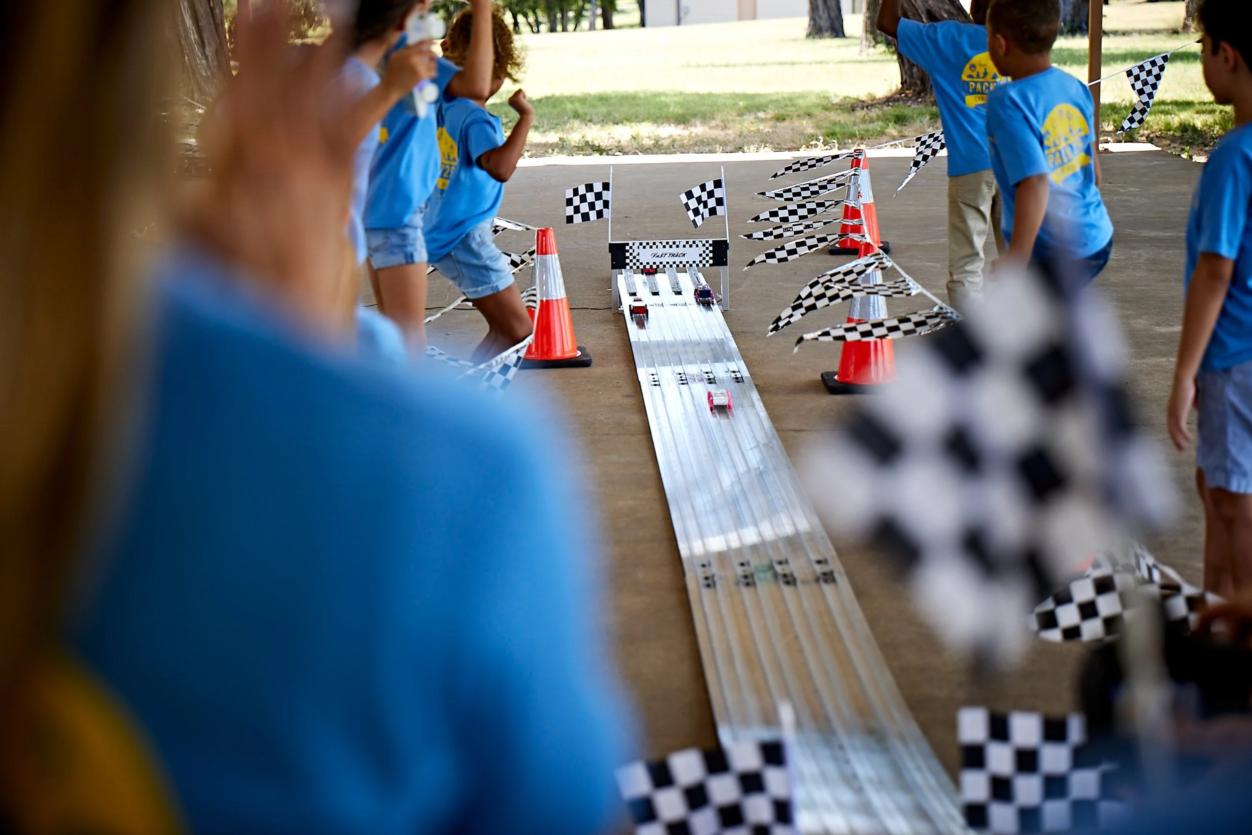 Scouts participating in a Pinewood Derby, with a race track set up outdoors and checkered flags decorating the scene.