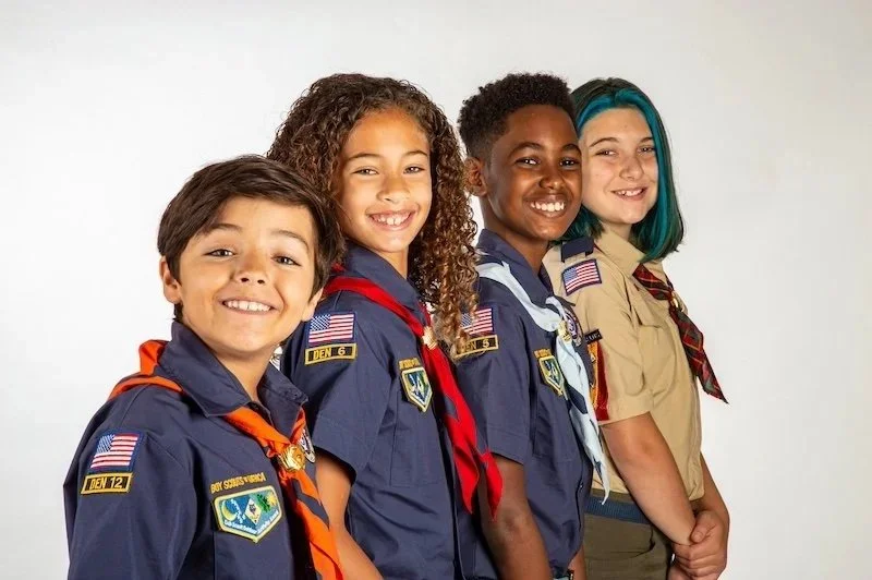 Four children dressed in scouting uniforms, standing side by side and smiling against a plain white background.