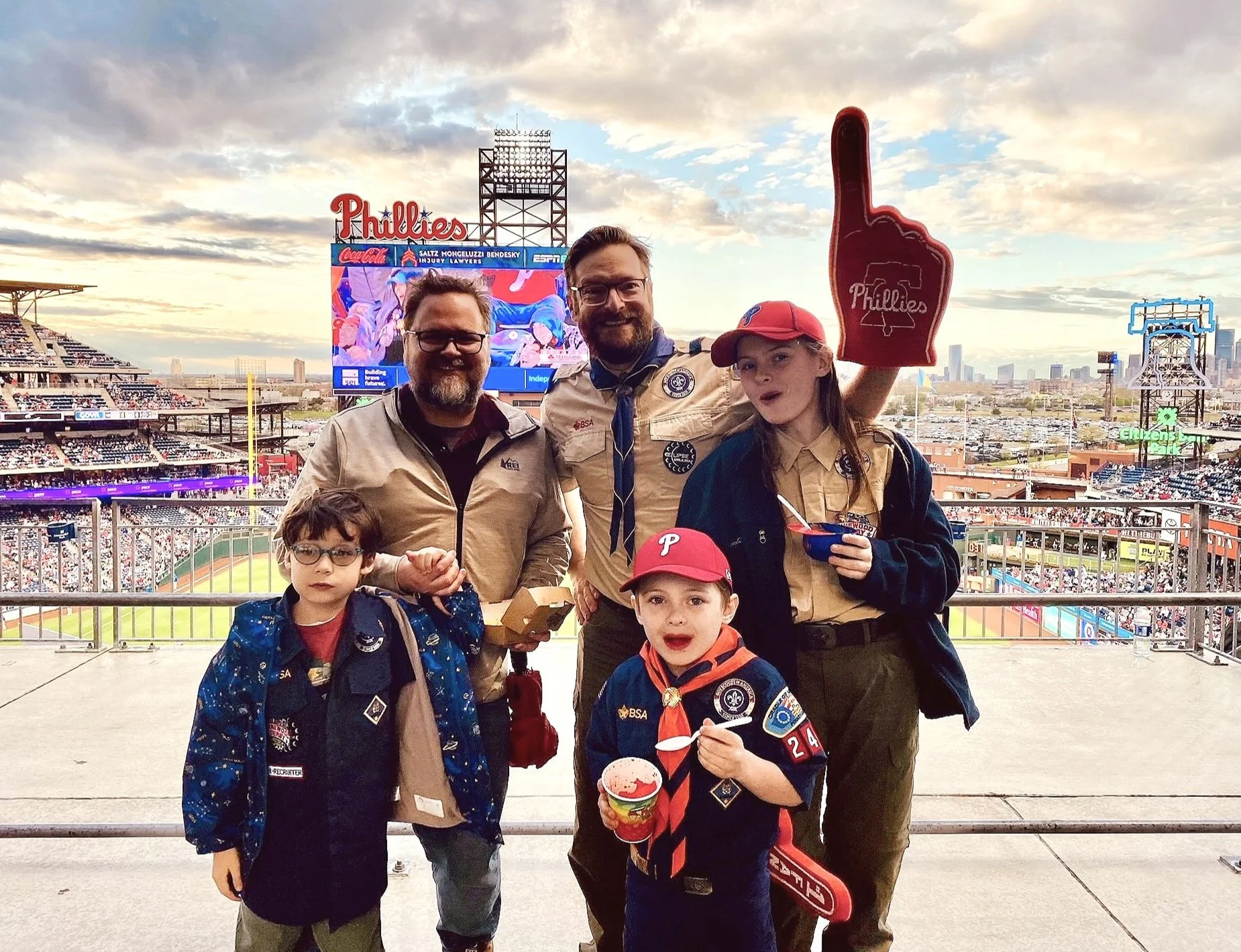 A group of Scouts and their families at a Phillies stadium with the city skyline in the background. They are wearing Phillies gear and holding snacks, smiling at the camera.