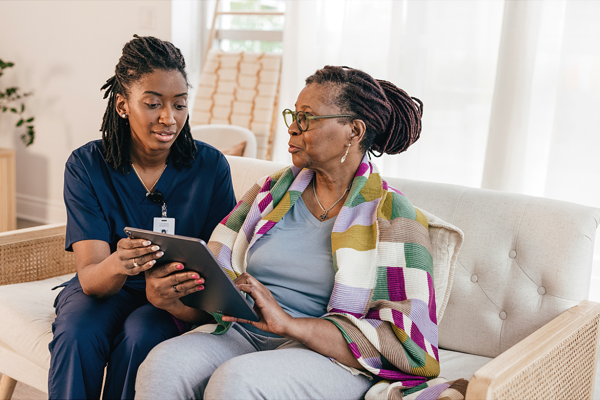 A nurse or caregiver and an elderly woman sitting on a beige sofa in a bright, airy room. The caregiver is showing something on a tablet to the elderly woman, who is wearing glasses and a colorful striped shawl.