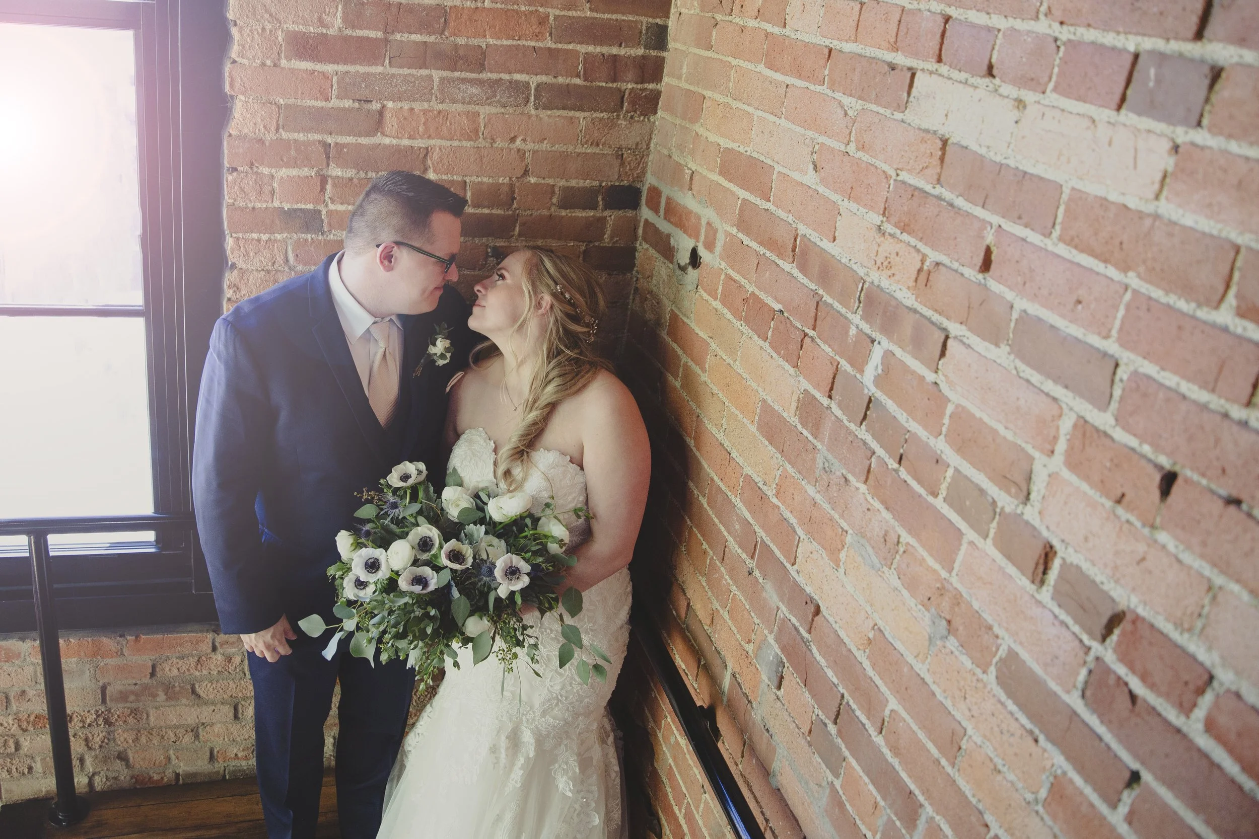 A bride and groom in wedding attire standing close together in front of a brick wall near a window, looking into each other's eyes.