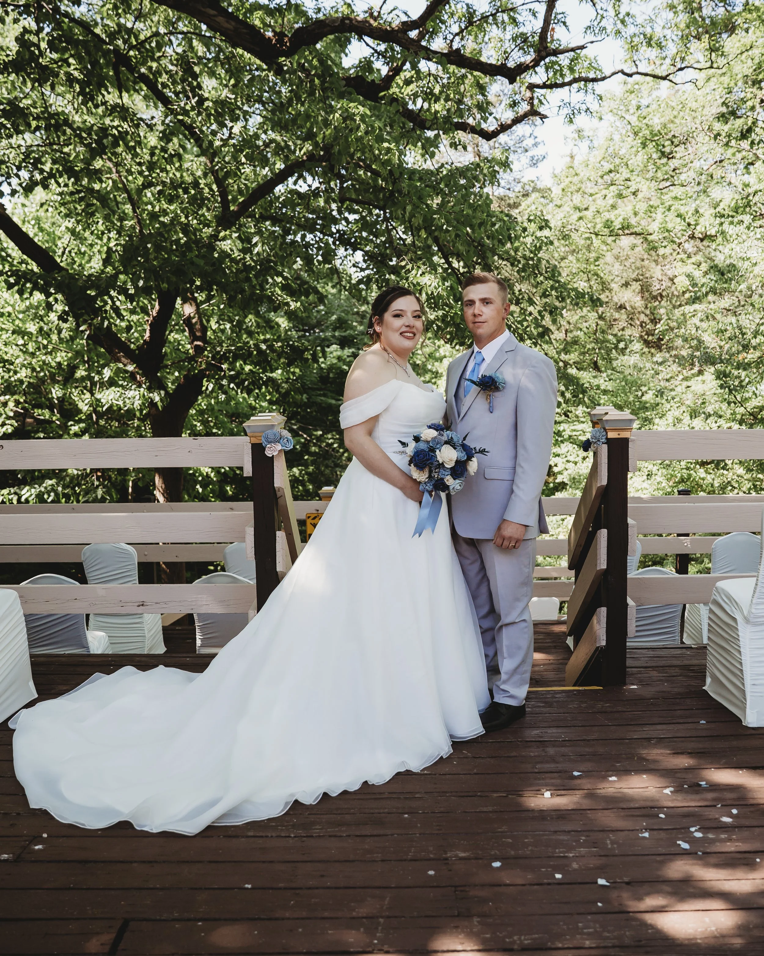 A bride and groom standing on a wooden platform outdoors, surrounded by green trees, dressed in wedding attire, holding a bouquet of blue and white flowers.
