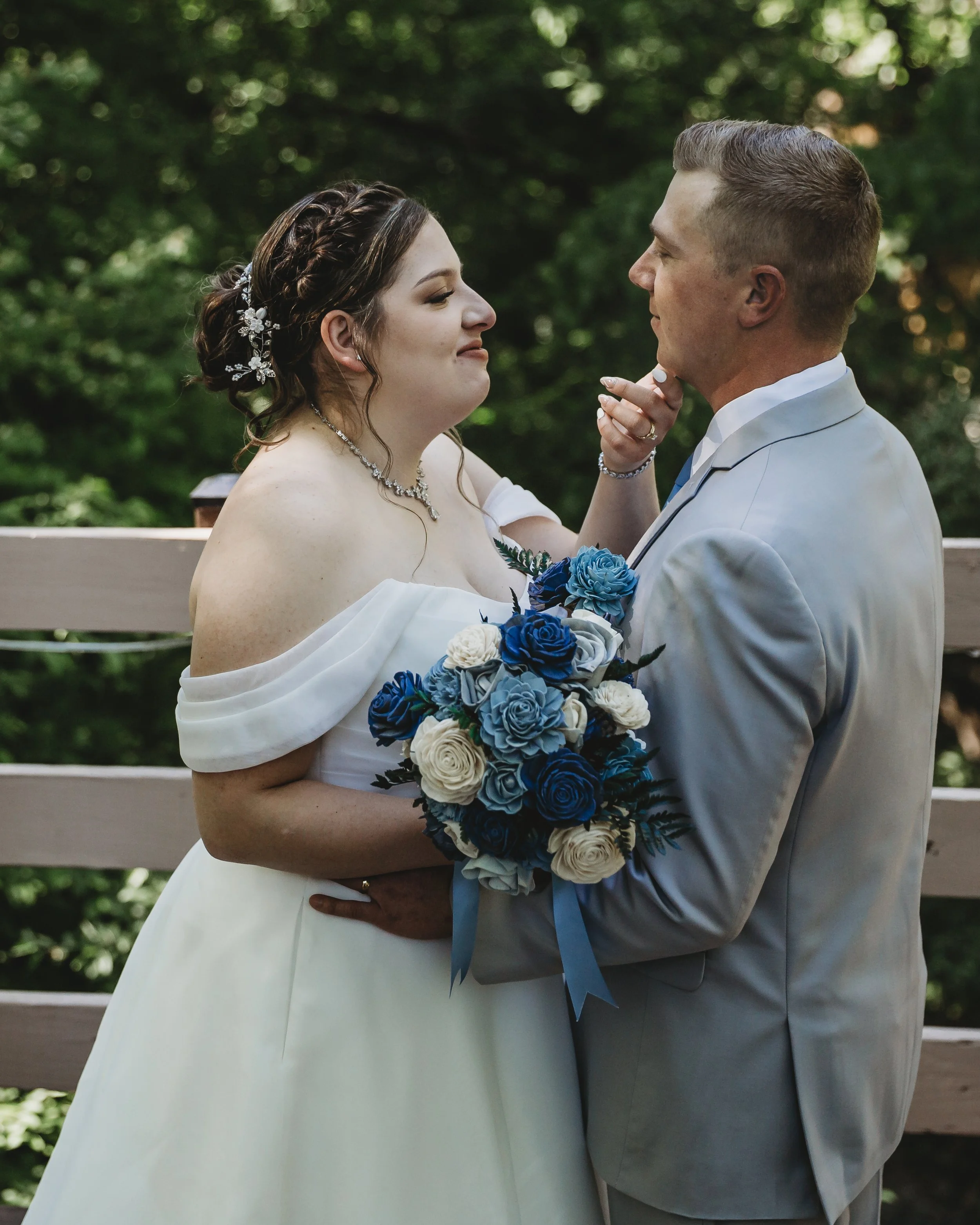 A bride and groom share an intimate moment outdoors, with the bride holding a bouquet of blue and white roses, and the groom gently touching her face as they look into each other's eyes.