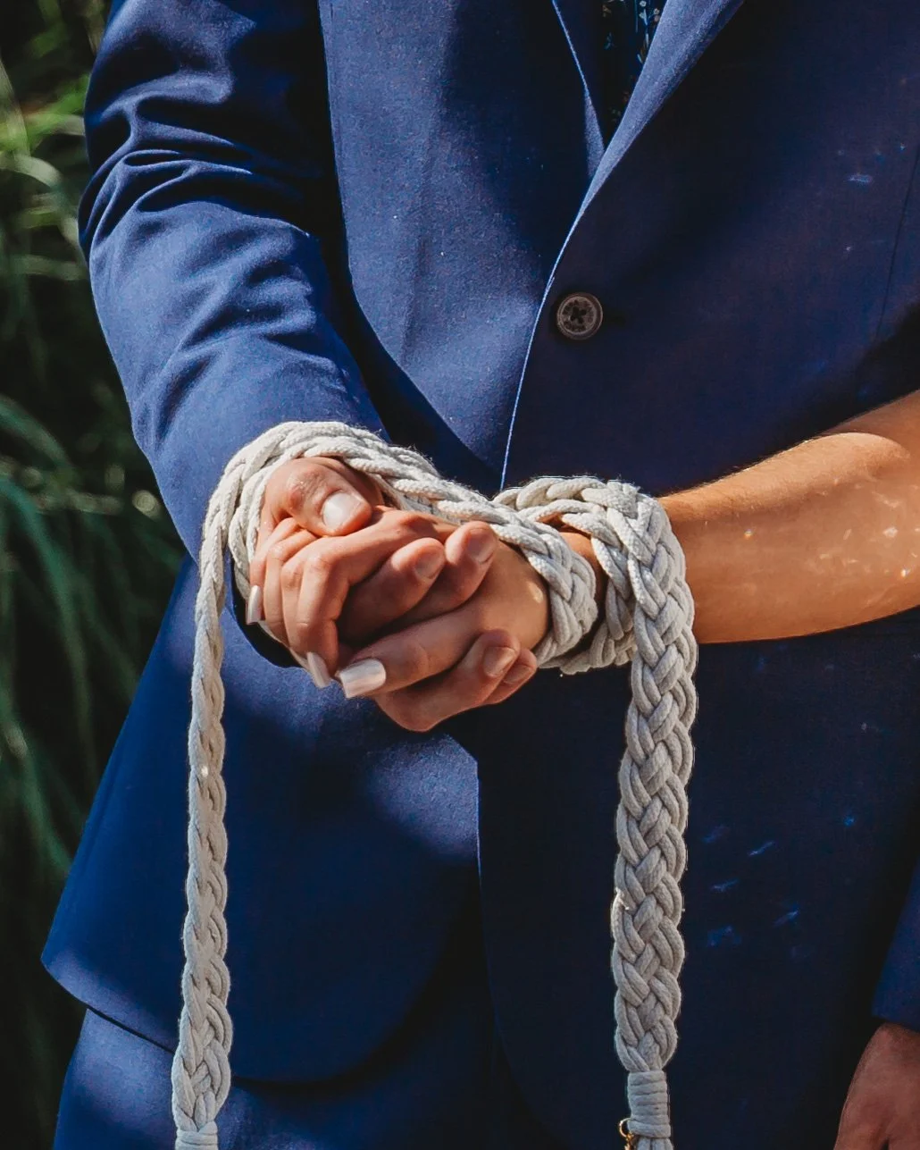 A person in a blue suit with hands bound together by a thick, beige rope, with the knots clearly visible.