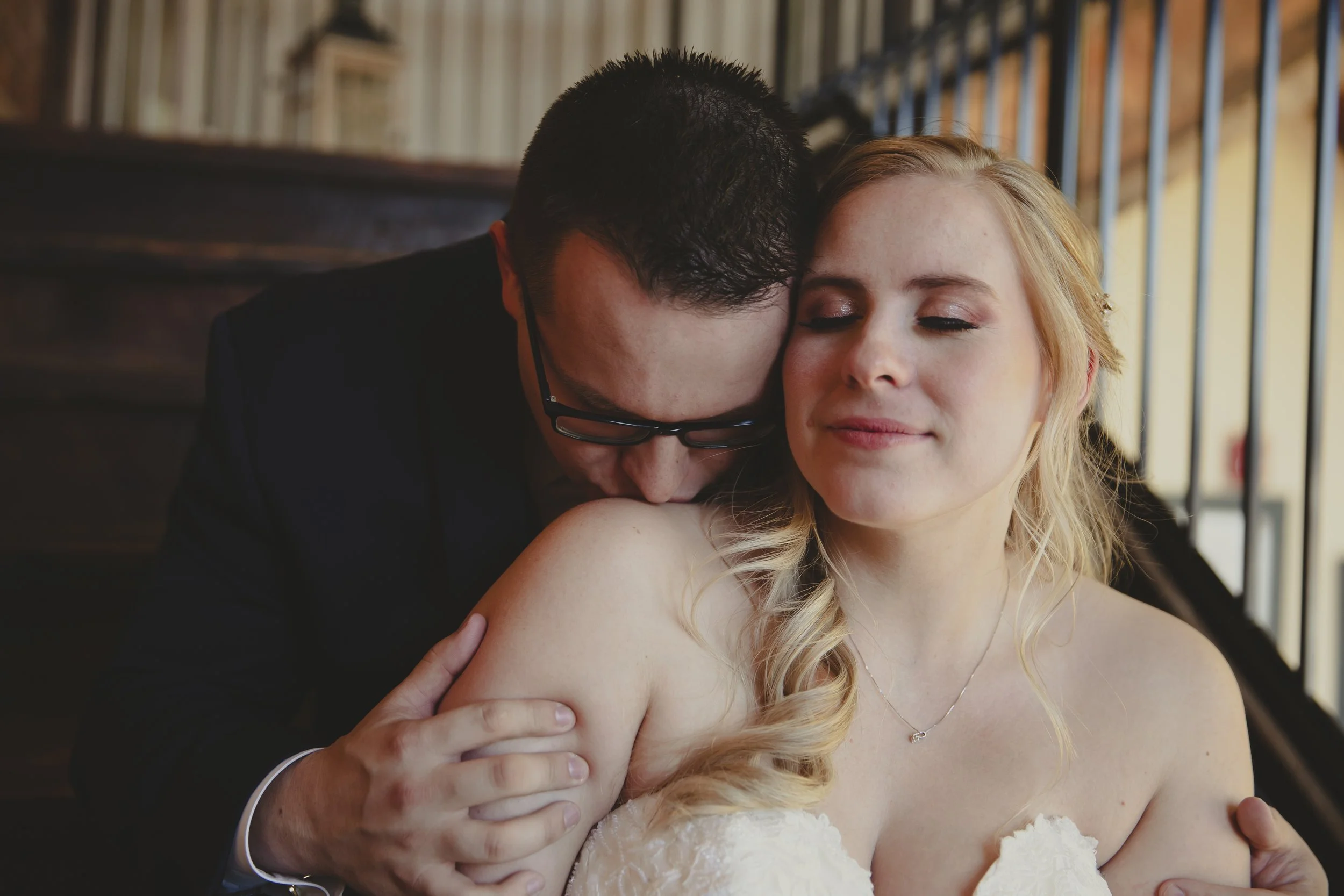 A man and woman sharing an intimate moment, with the man gently holding the woman and kissing her shoulder, in a warmly lit indoor setting.