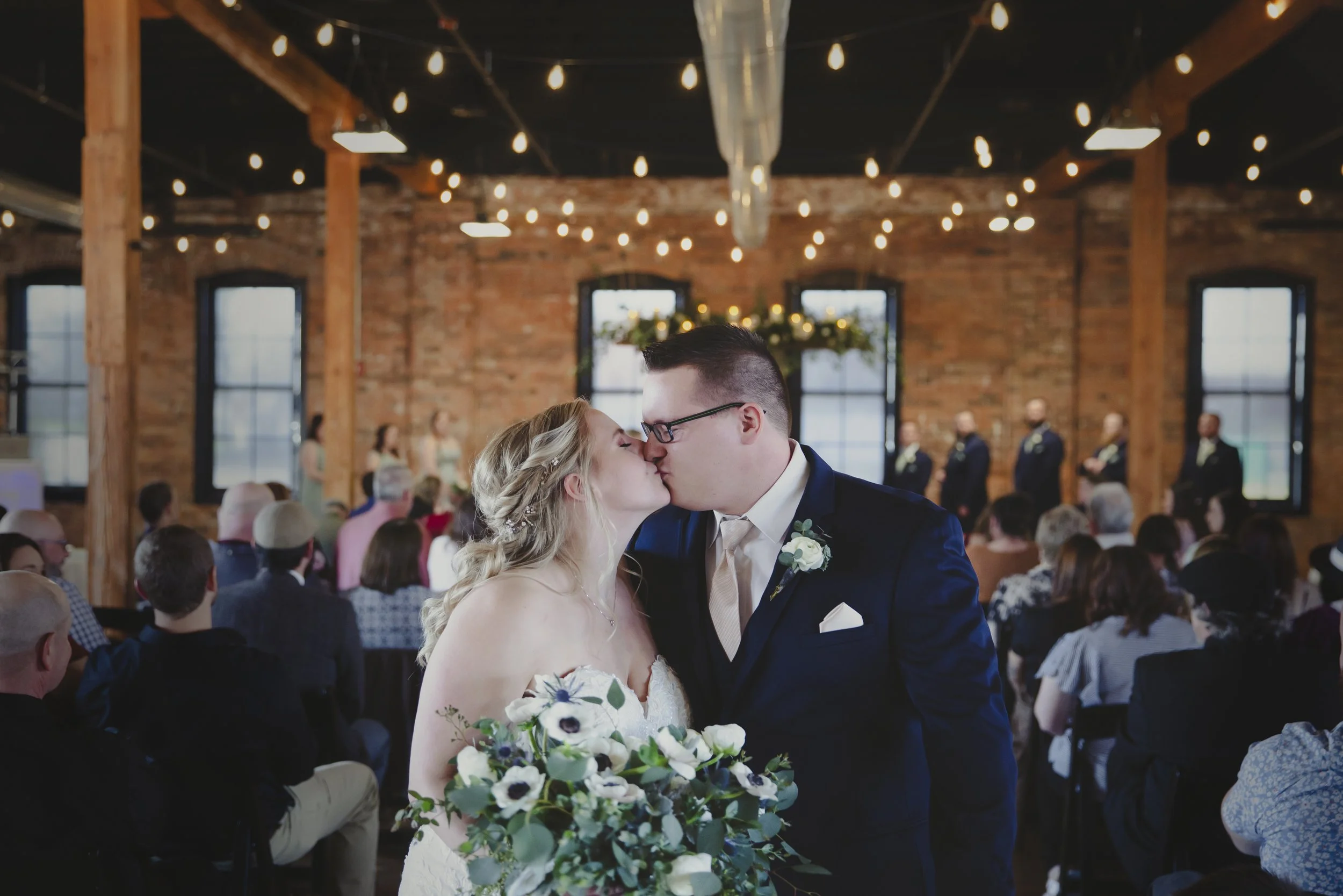 A bride and groom share a kiss during their wedding ceremony in a rustic indoor venue with exposed brick walls, large windows, and string lights hanging from the ceiling.