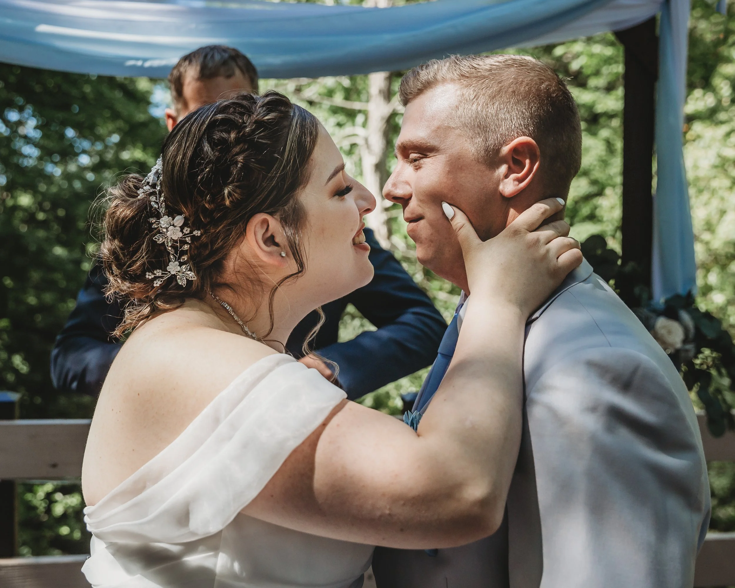 A bride and groom are close together during their wedding ceremony outdoors, smiling and gazing at each other with greenery in the background.