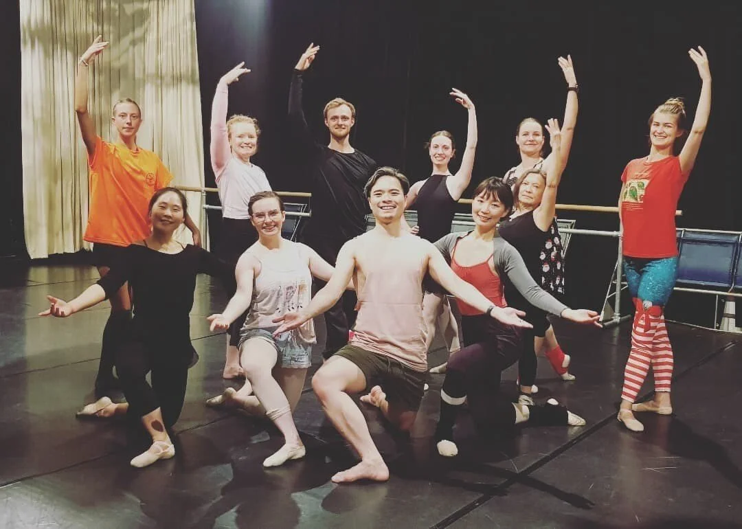 Ballet dancers posing in a studio with a black stage and beige curtains, some standing and some kneeling in ballet poses.