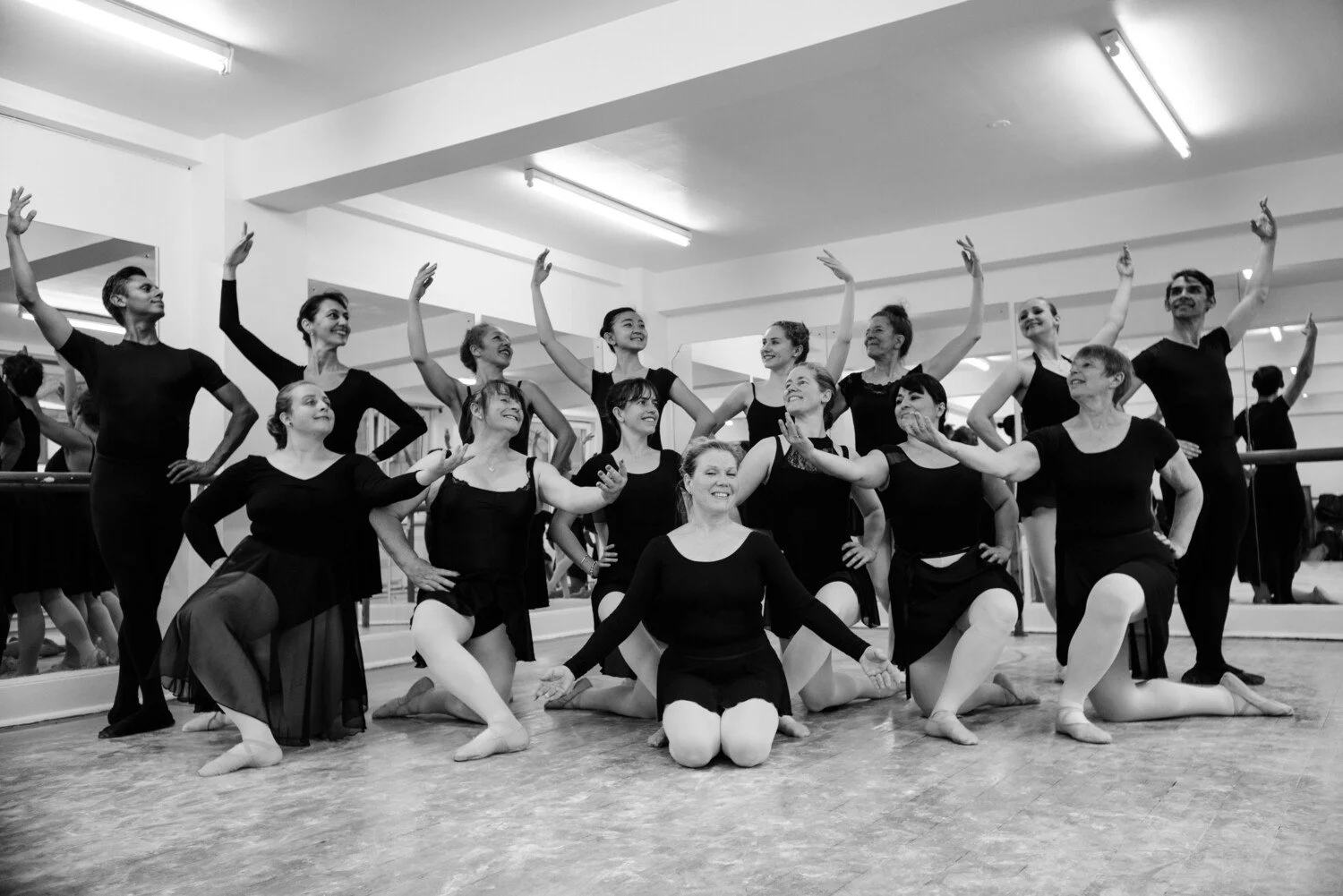 Group of ballet dancers in black costumes posing in a dance studio, some kneeling in front and some standing behind, smiling and making expressive gestures.