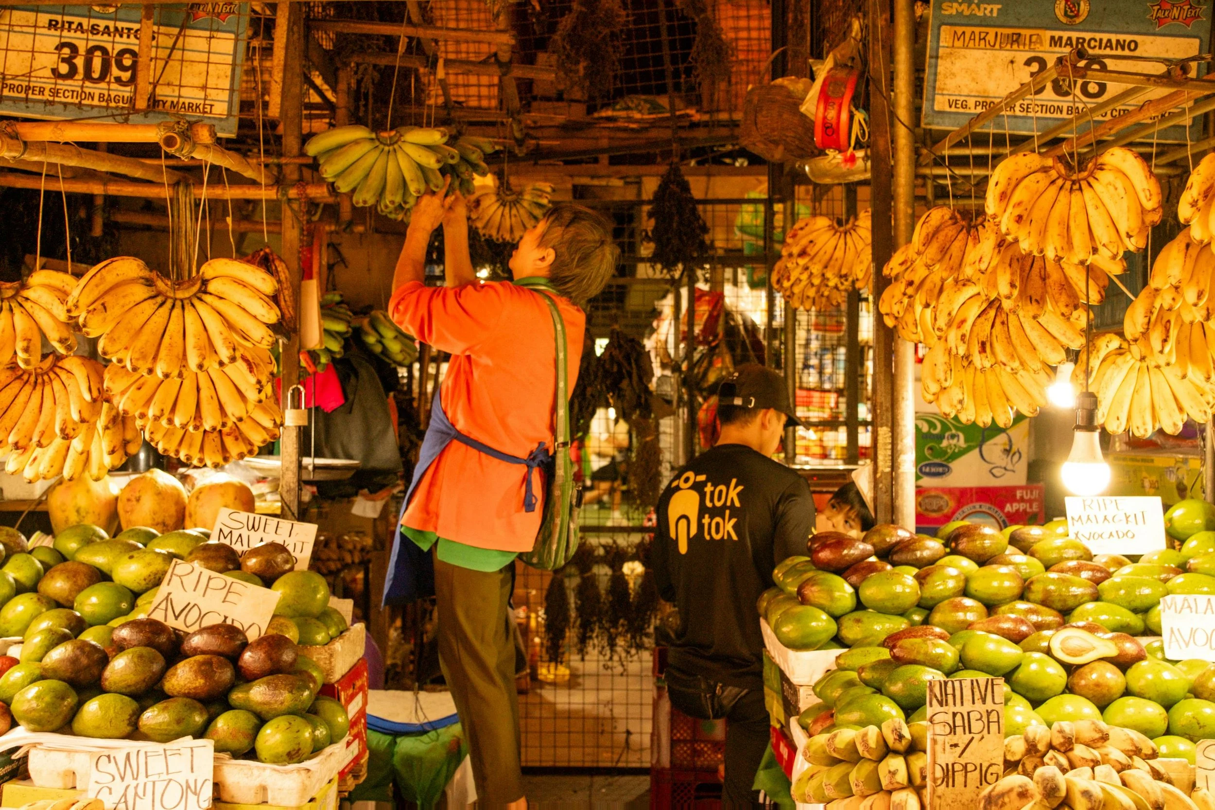 A market stall selling bananas, avocados, and other fruits, with vendors and a customer shopping, illuminated by warm lighting.