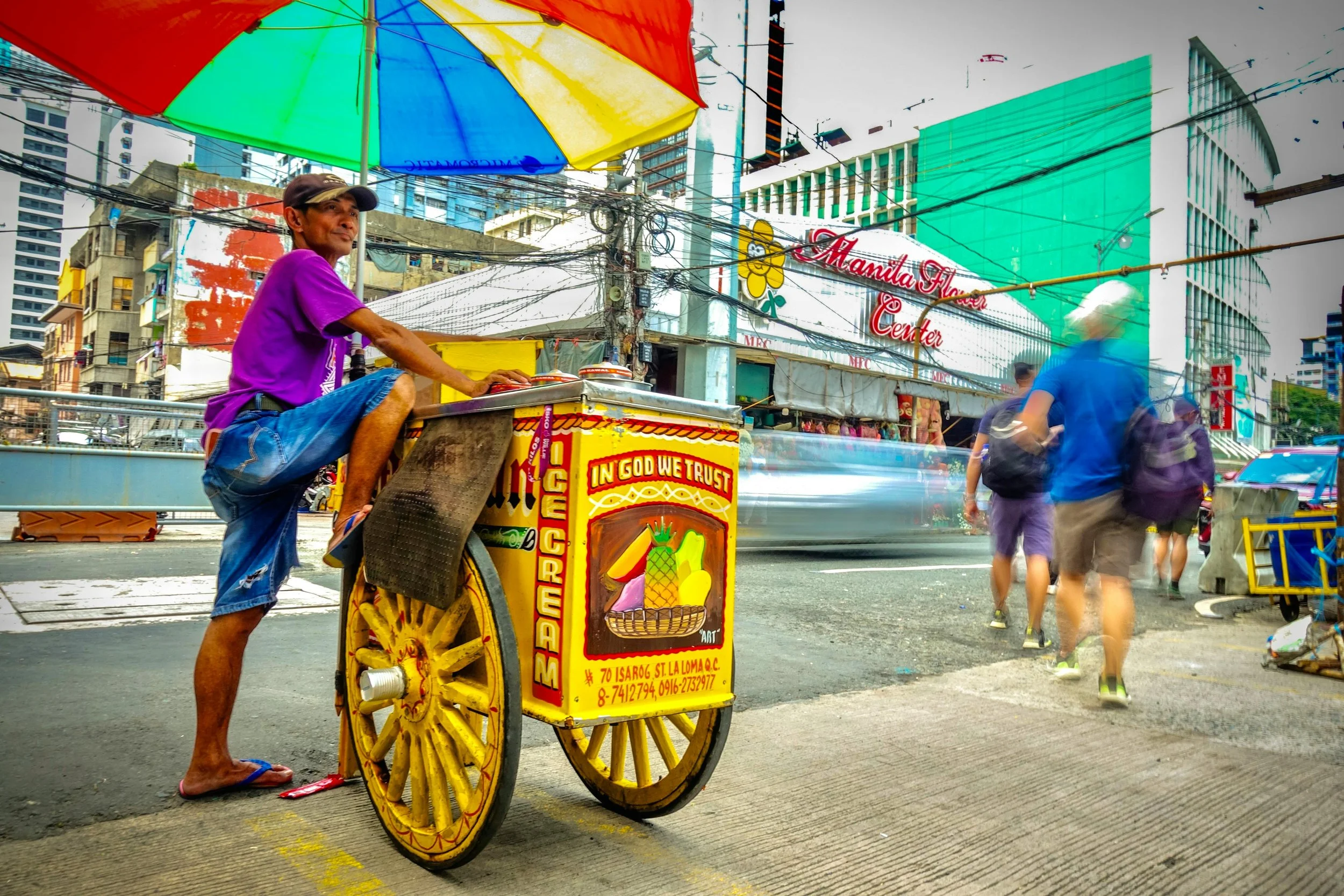 A street scene in Manila with a man in a purple shirt and blue shorts selling food from a colorful cart under a multicolored umbrella. Blurred pedestrians walk by, and a Manila Flower Center sign is visible in the background.