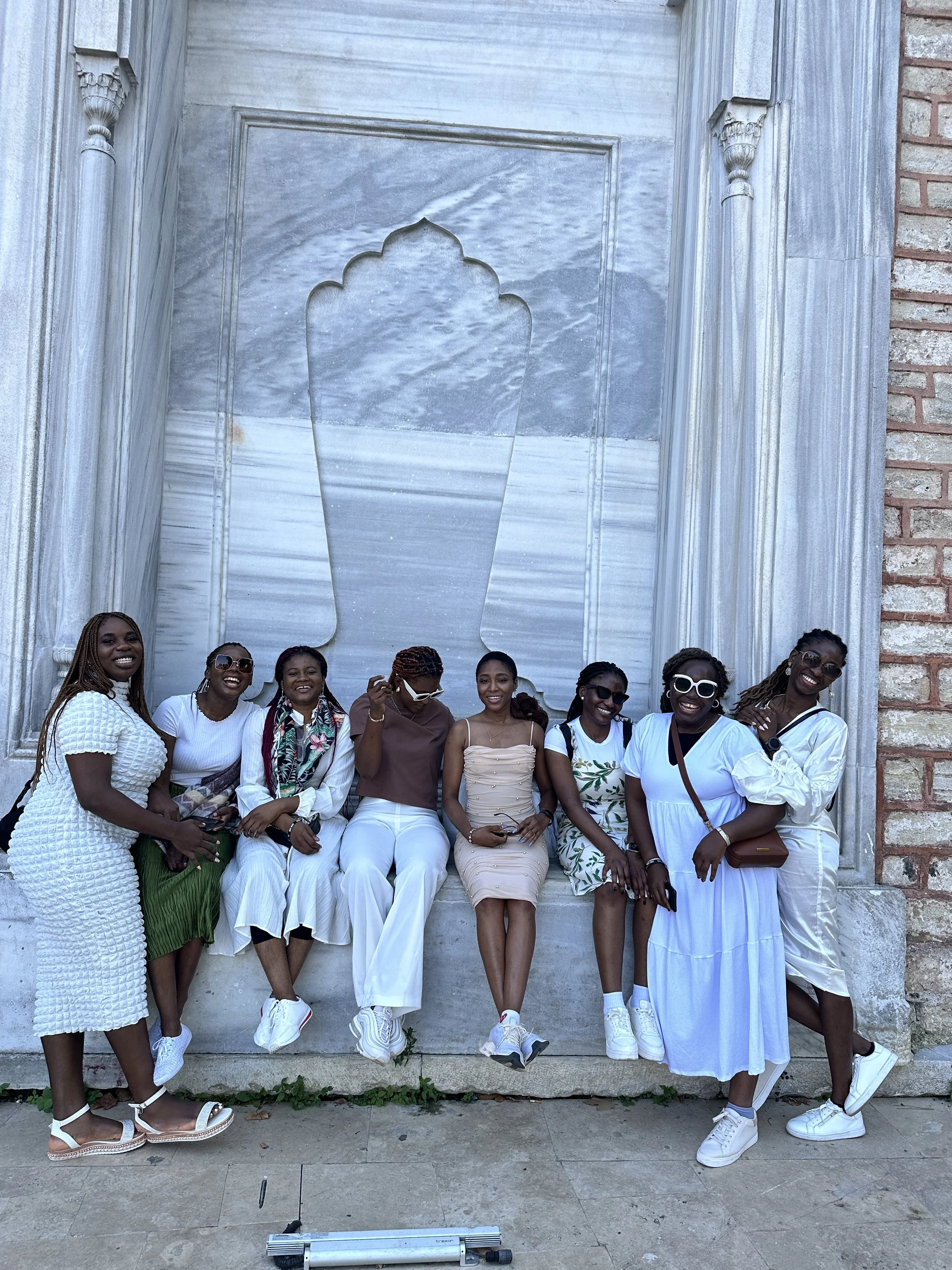 A group of nine women posing and smiling in front of a large water feature with a decorative gate at a tourist site, all dressed in light-colored clothing.