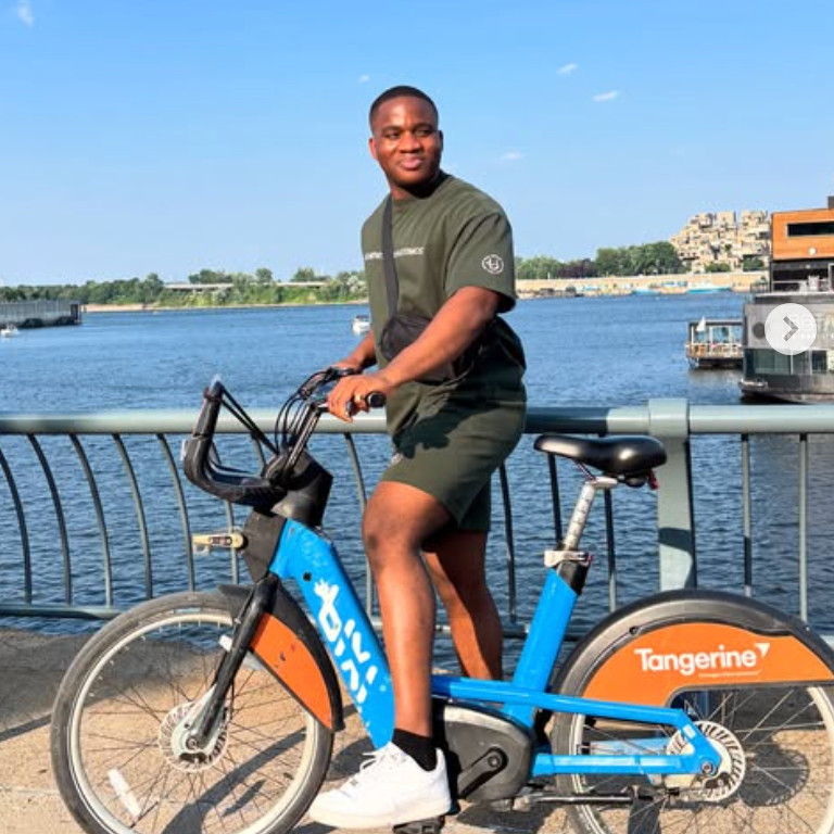 Young man riding a blue and orange bicycle near a river with buildings in the background on a sunny day.