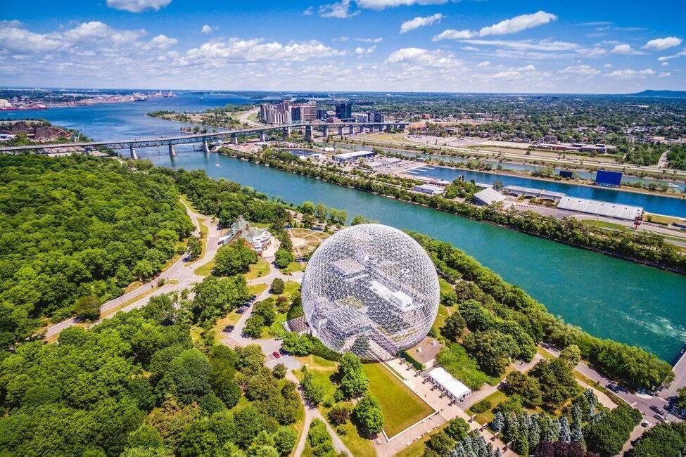 Aerial view of a city landscape featuring a large glass sphere structure within a park, with a river, bridges, and urban buildings in the background.