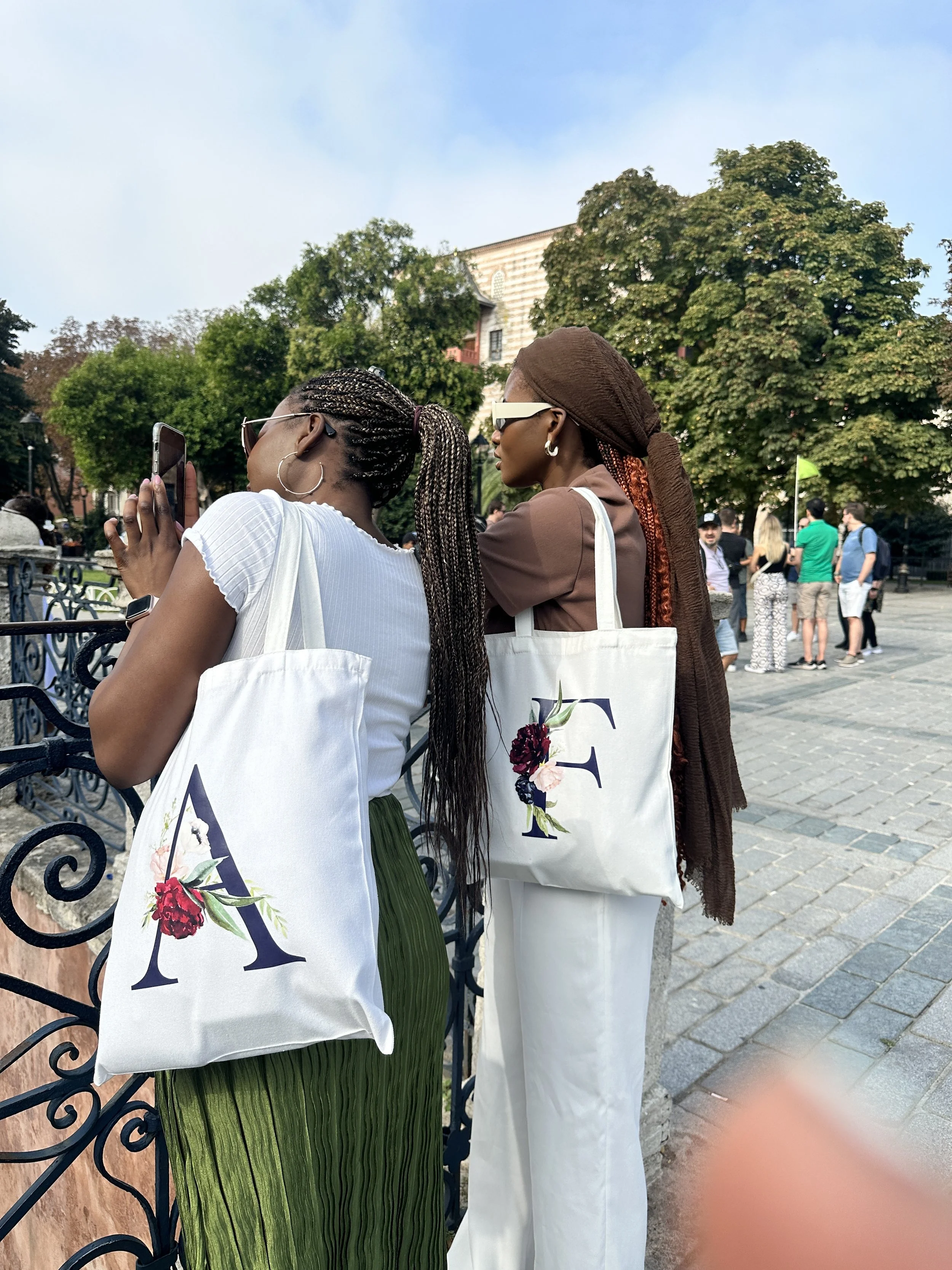 Two women standing near a black wrought iron fence, one taking a photo with her smartphone. Both women carry white tote bags with large styled letter 'F' and floral designs. There are trees and a group of people in the background on a city street.