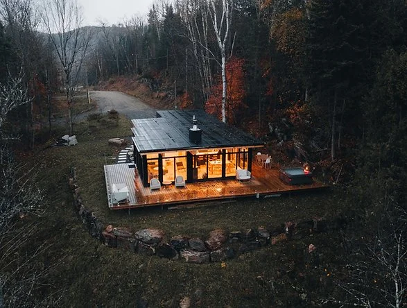 A modern house with large glass windows and a wooden deck is surrounded by trees in a wooded area, with a winding driveway nearby.