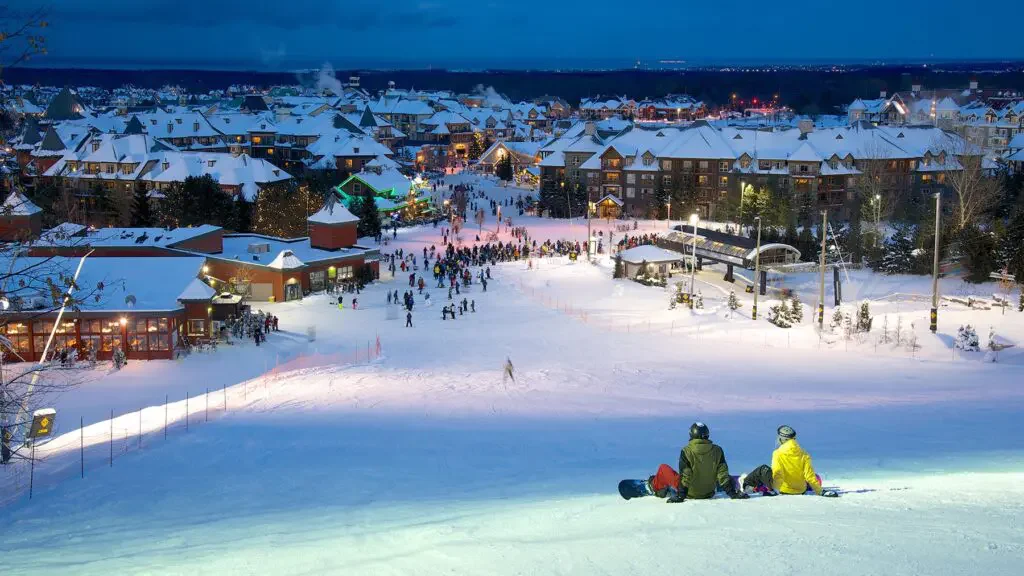 People enjoying a snowy evening at a ski resort, with skiers and visitors on the slopes and in the town. Two individuals sit on the snow wearing ski gear, overlooking the scene.