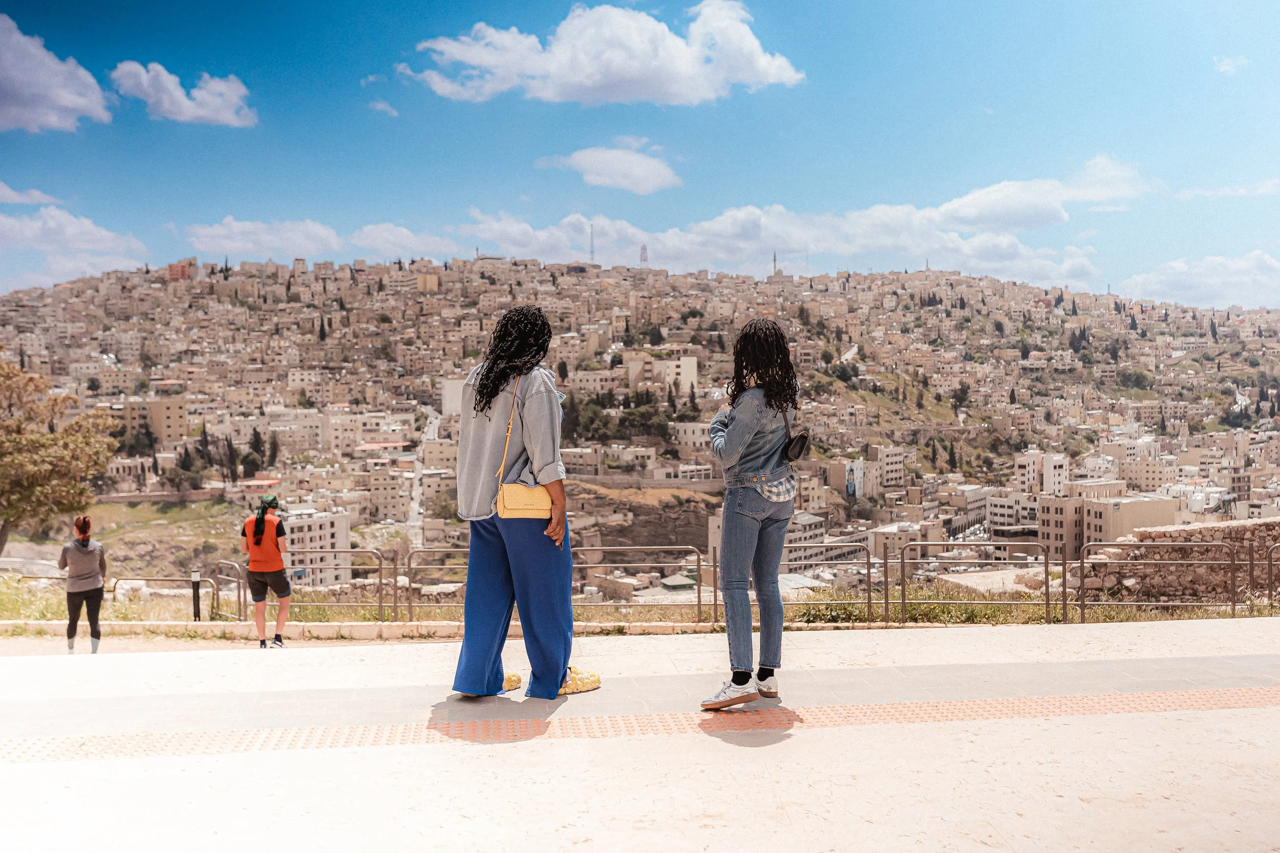 Two women and two men stand on a viewpoint overlooking a hilly cityscape with many beige buildings under a partly cloudy sky.