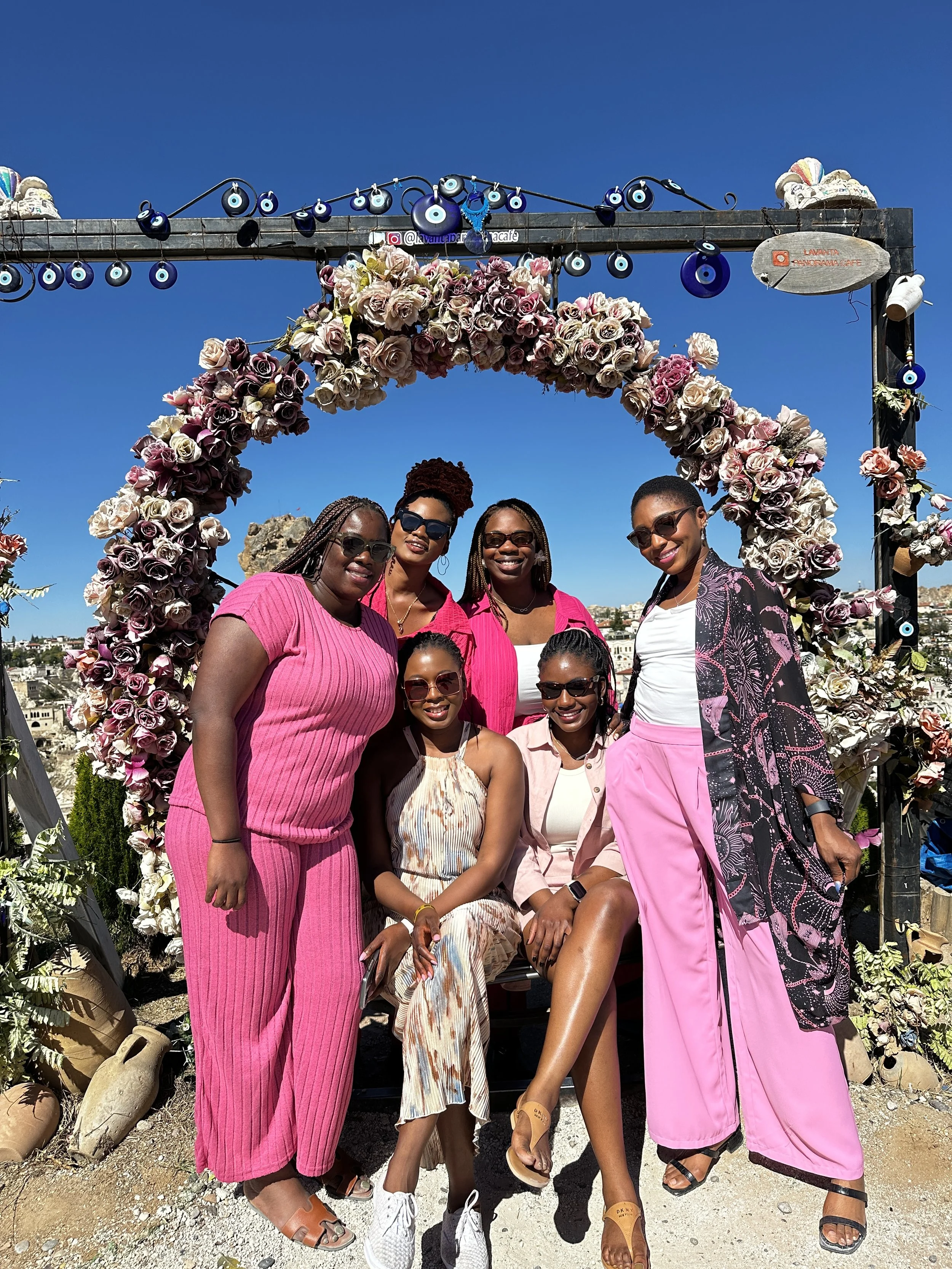 A group of six women with dark skin, wearing stylish pink, white, and beige outfits, standing and sitting in front of a heart-shaped floral arch decorated with pink and white roses on a bright sunny day.