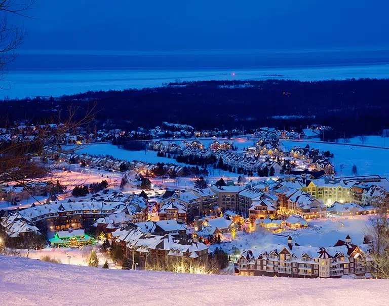 Nighttime view of a snow-covered town with illuminated buildings and houses, surrounded by a snowy landscape and distant dark hills.