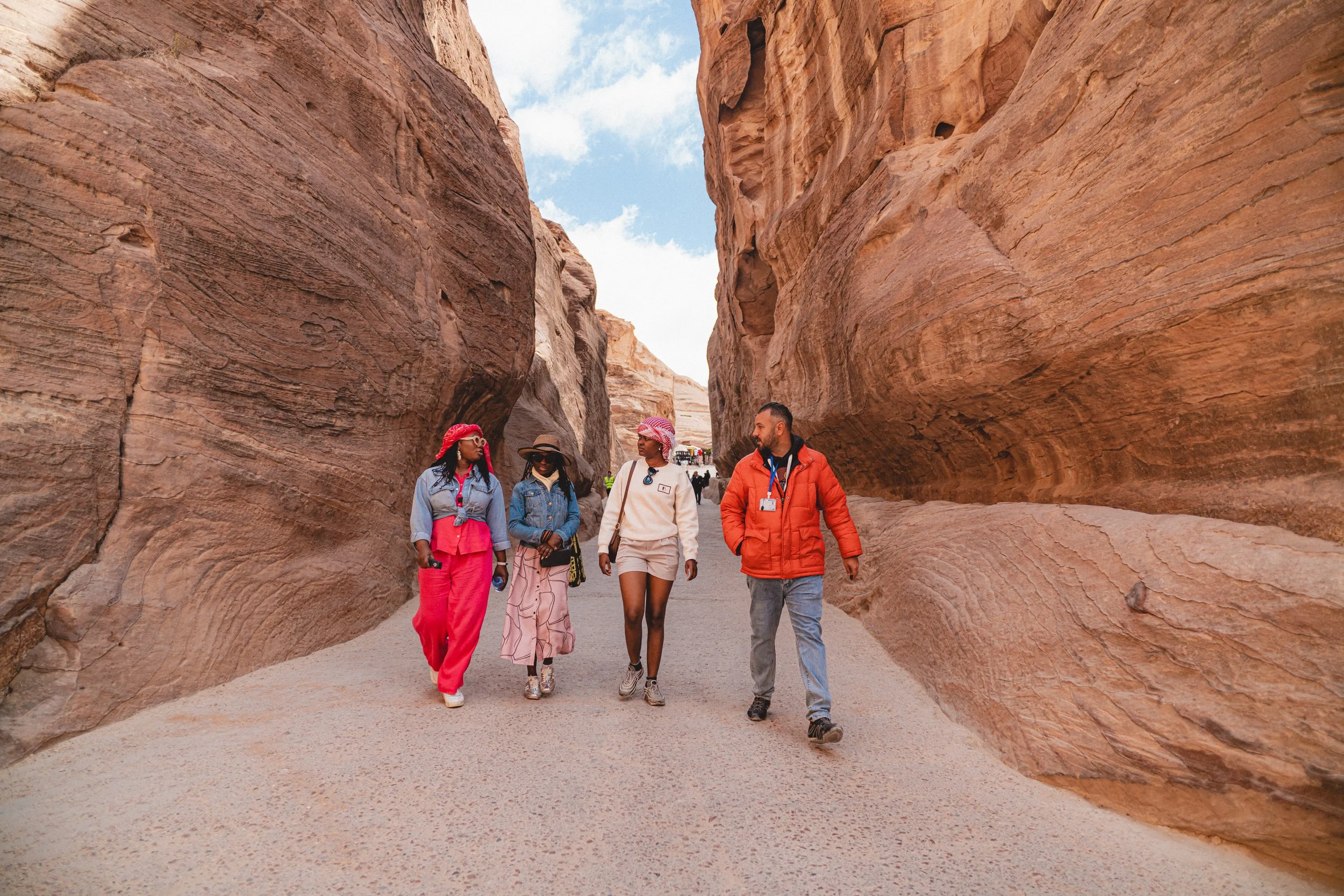 Four people walking through a narrow canyon with tall, reddish-brown rock walls on either side.