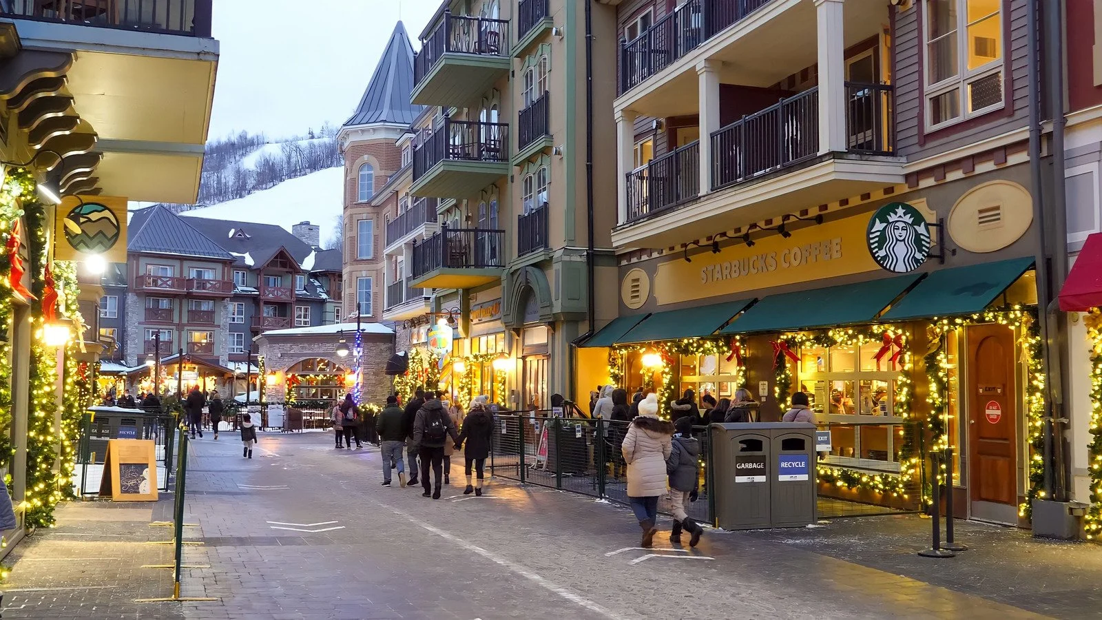 A lively street scene in a mountain town during the holiday season, featuring a Starbucks coffee shop decorated with Christmas lights and ornaments, with people walking and shopping, snowy mountain in the background, and festive decorations along the street.