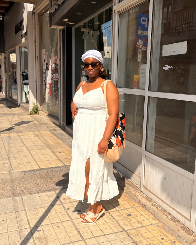 A woman in a white dress, white headwrap, and sunglasses standing on a city sidewalk in front of glass storefronts with posters and signs.