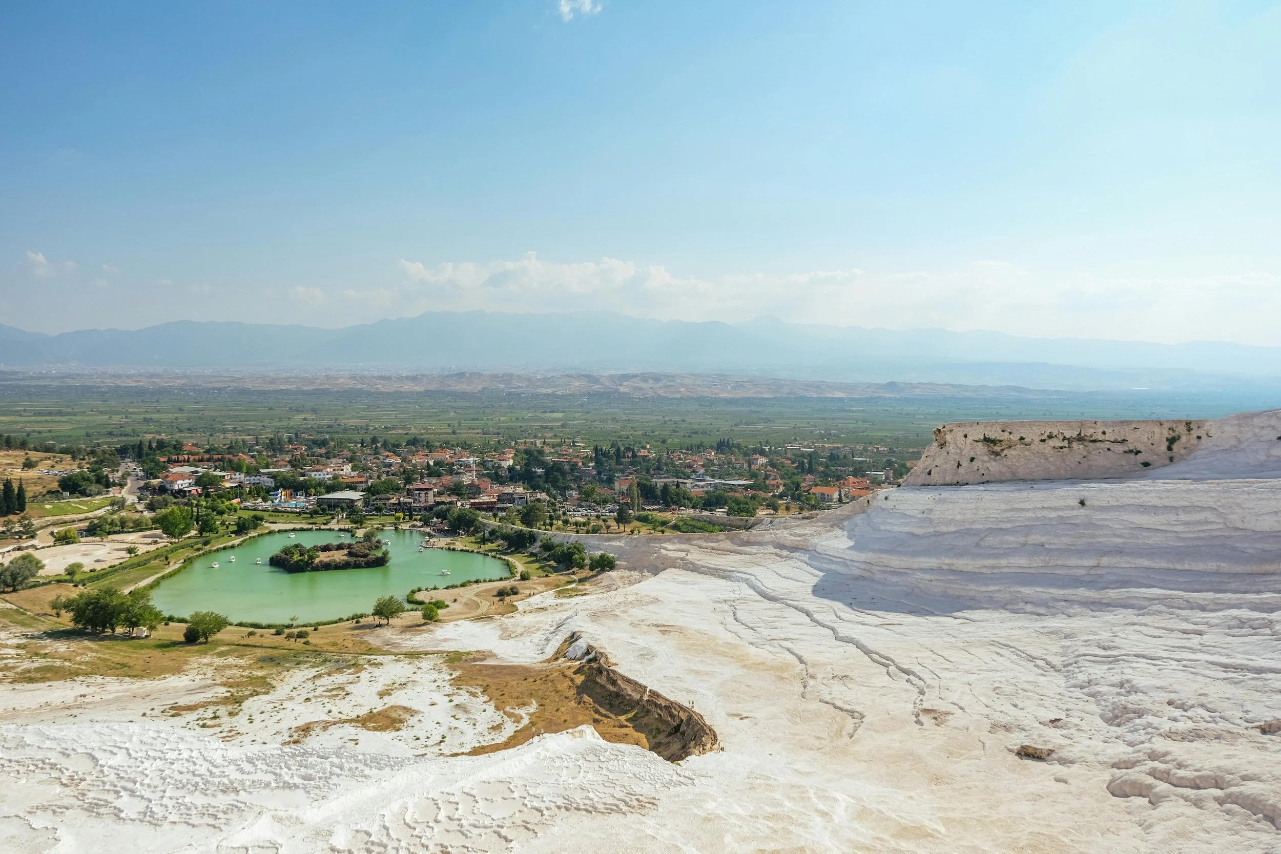 View of Pamukkale's white terraces and mineral pools with a small green lake and a town in the distance under a blue sky.
