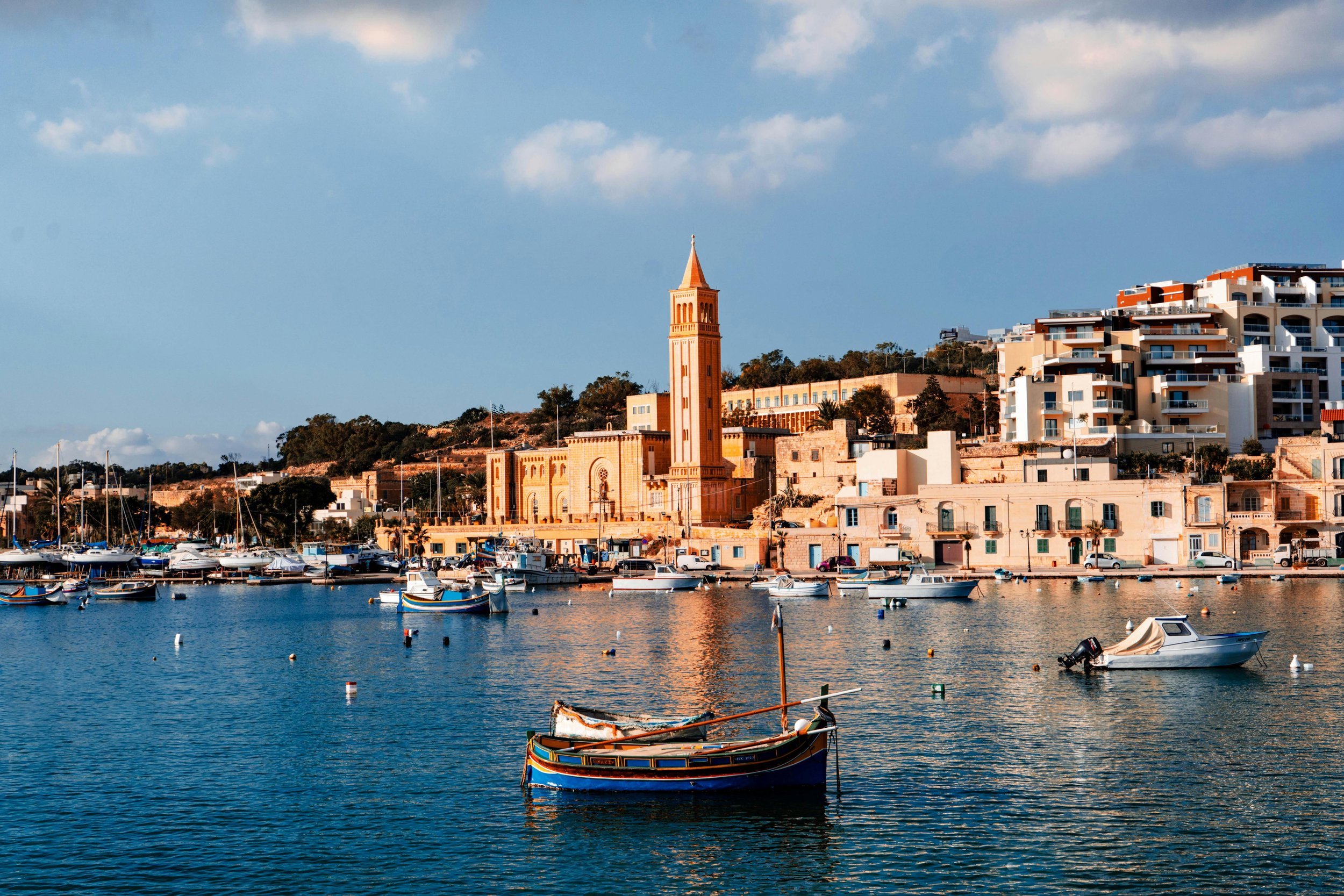 A harbor with several boats, colorful buildings, and a tall clock tower against a partly cloudy sky.