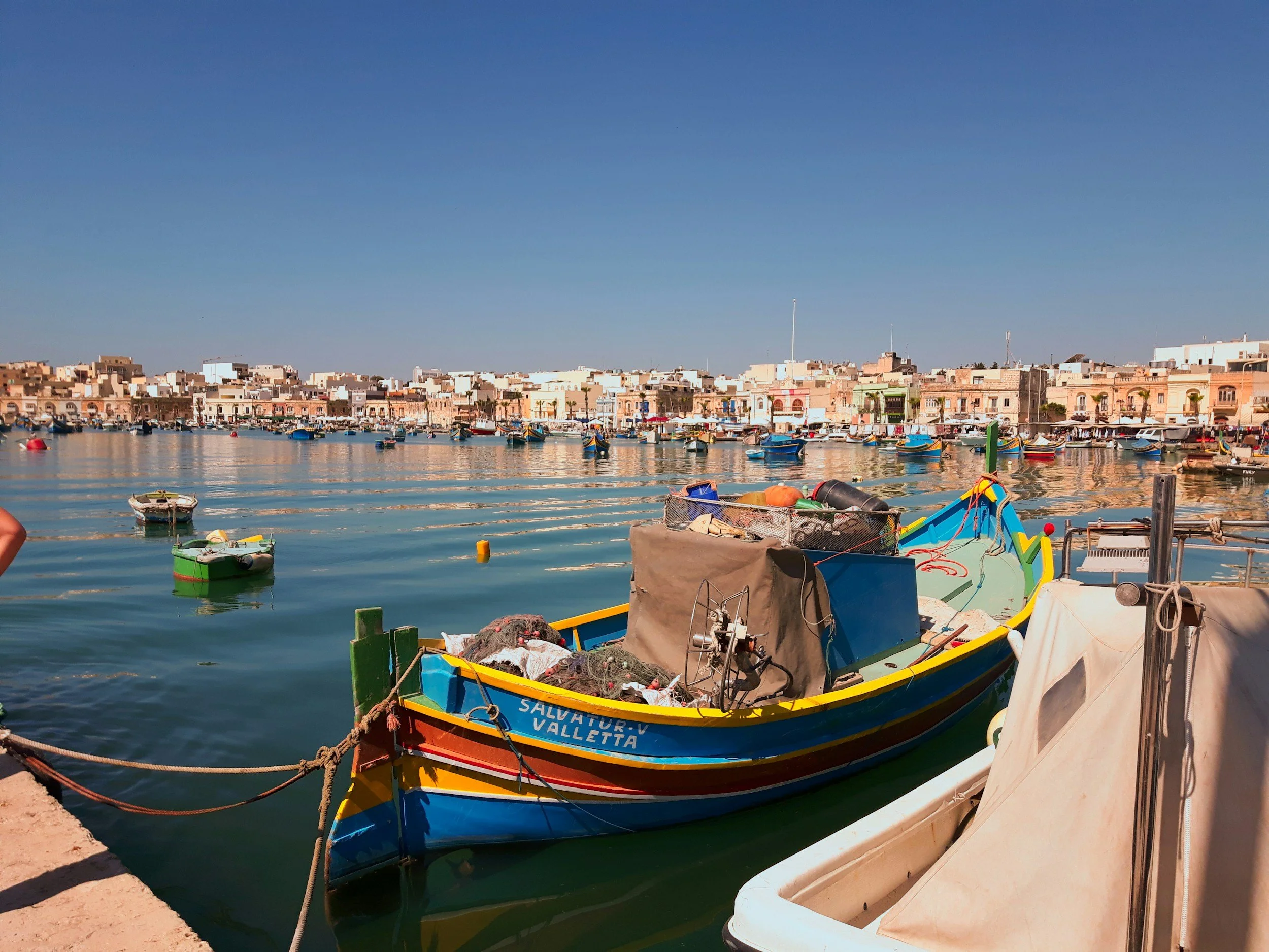 Colorful boats docked along a waterfront with a town of pastel-colored buildings and a clear blue sky in the background.