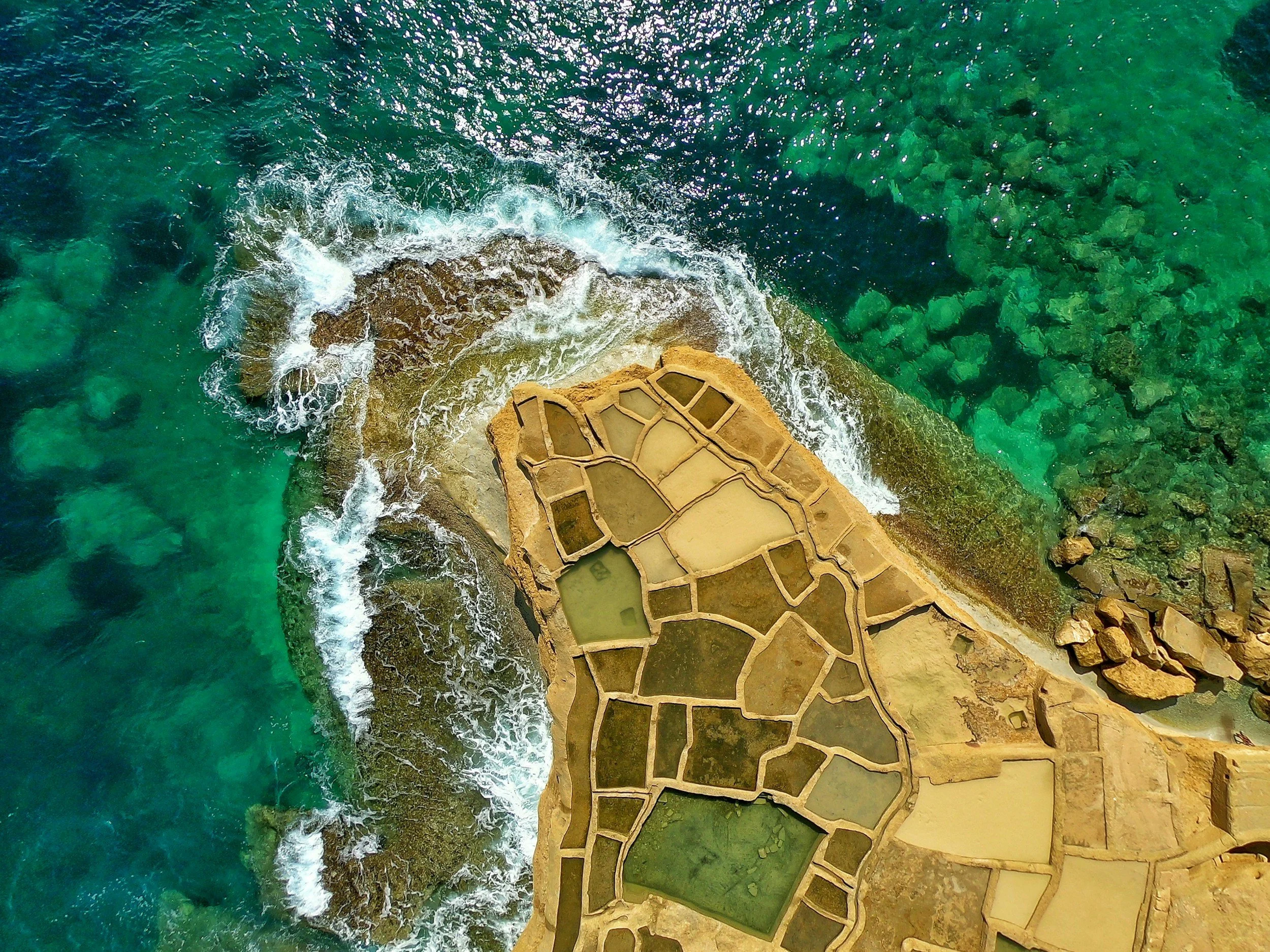 Aerial view of a coastline showing rocks, sea waves, and salt evaporation ponds with various shades of green and yellow.