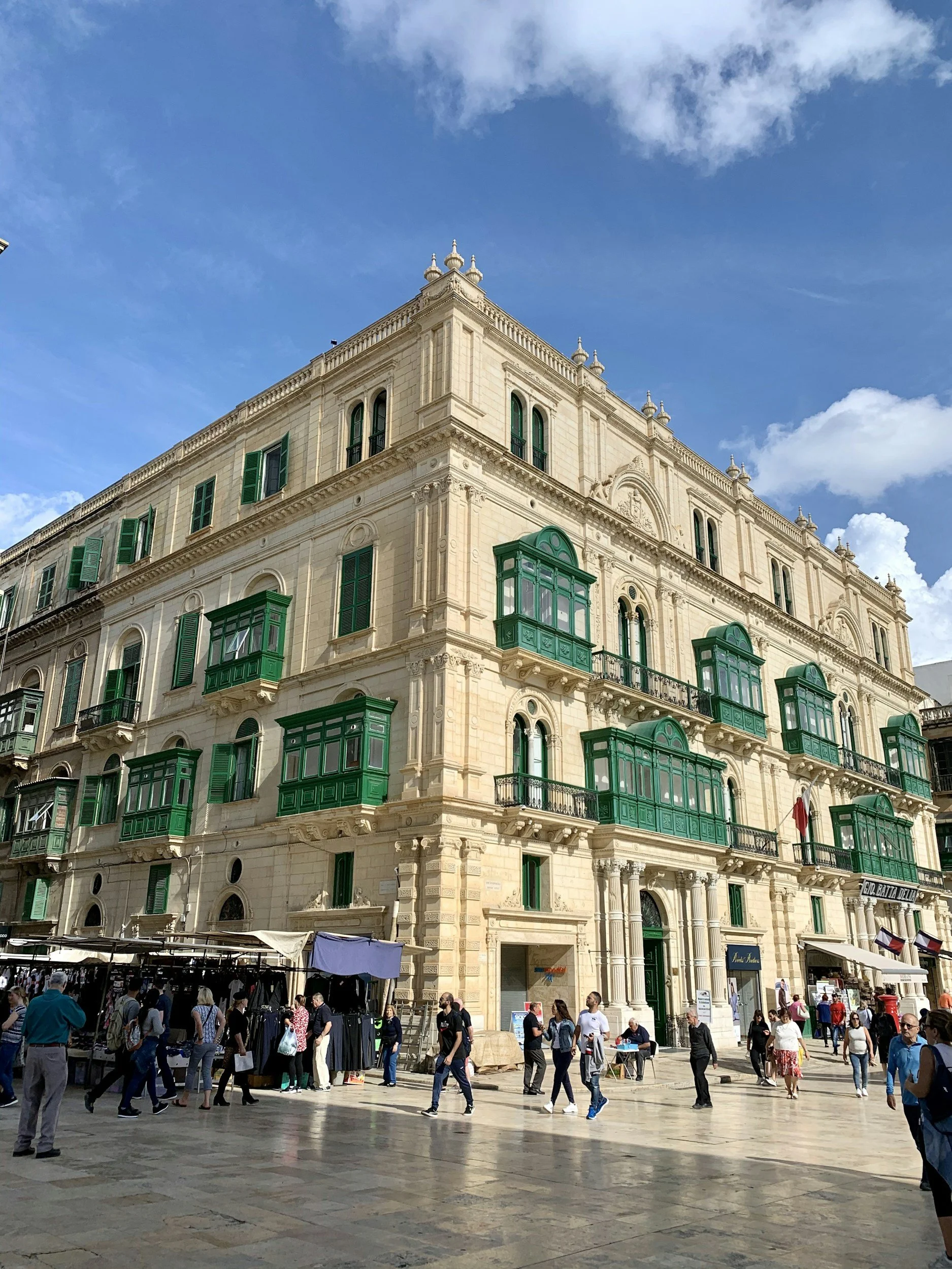 Historic building with beige stone exterior, multiple green balconies, and green window shutters, situated on a busy street with pedestrians and market stalls.