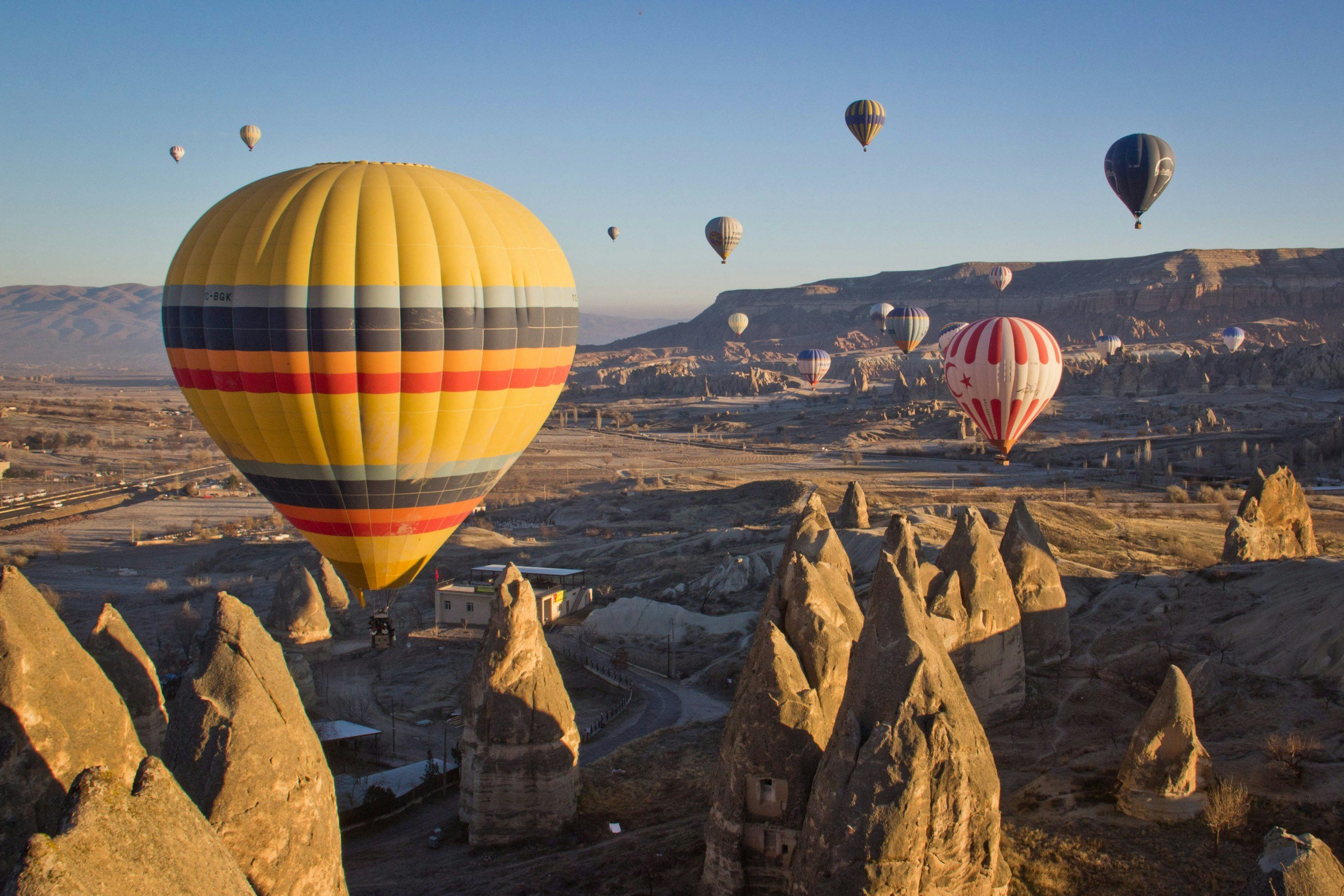 Multiple colorful hot air balloons floating over rocky landscape and fairy chimney formations during sunrise or sunset.