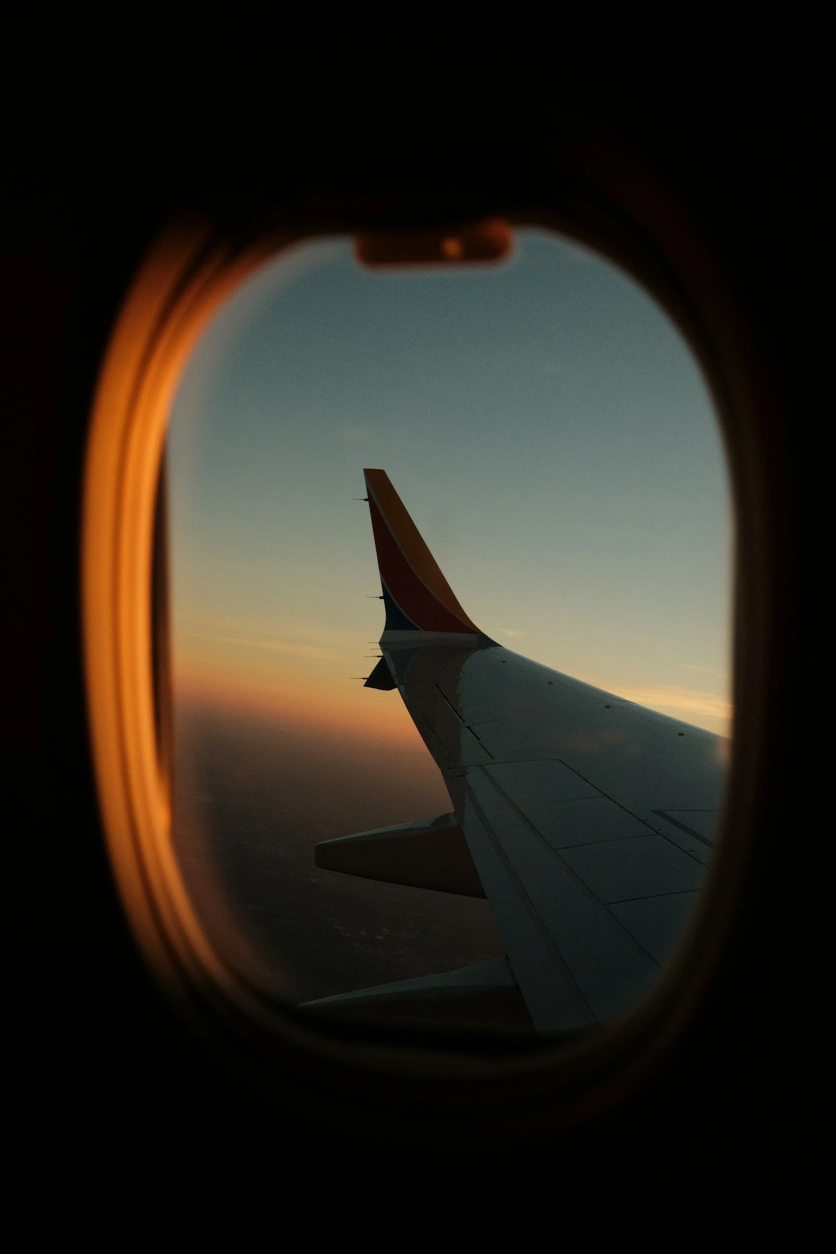 View of an airplane wing through a window during sunset or sunrise, with the sky in shades of orange and blue.