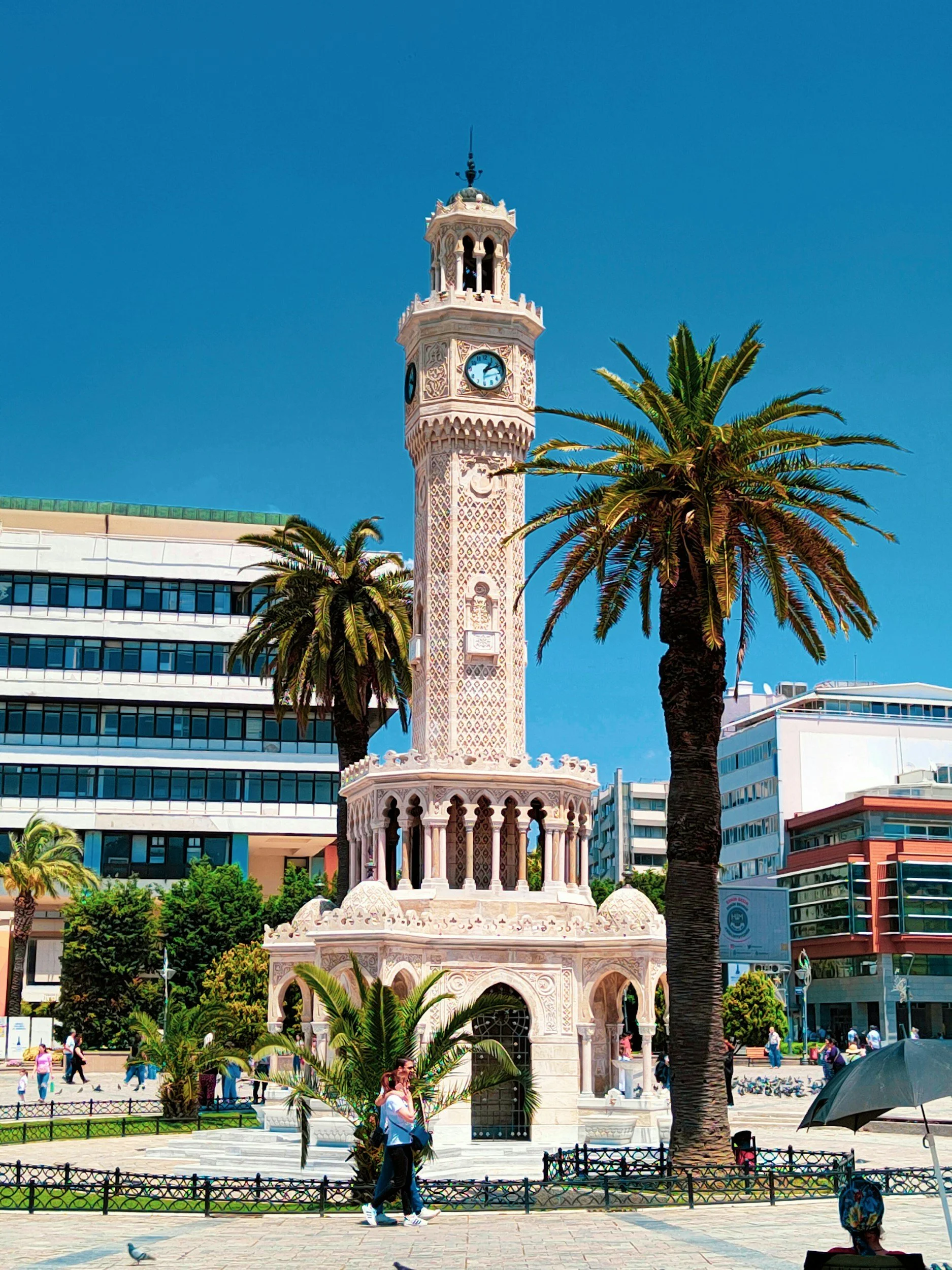 A historic clock tower with intricate carvings, surrounded by palm trees, in an urban park with modern buildings in the background on a sunny day.